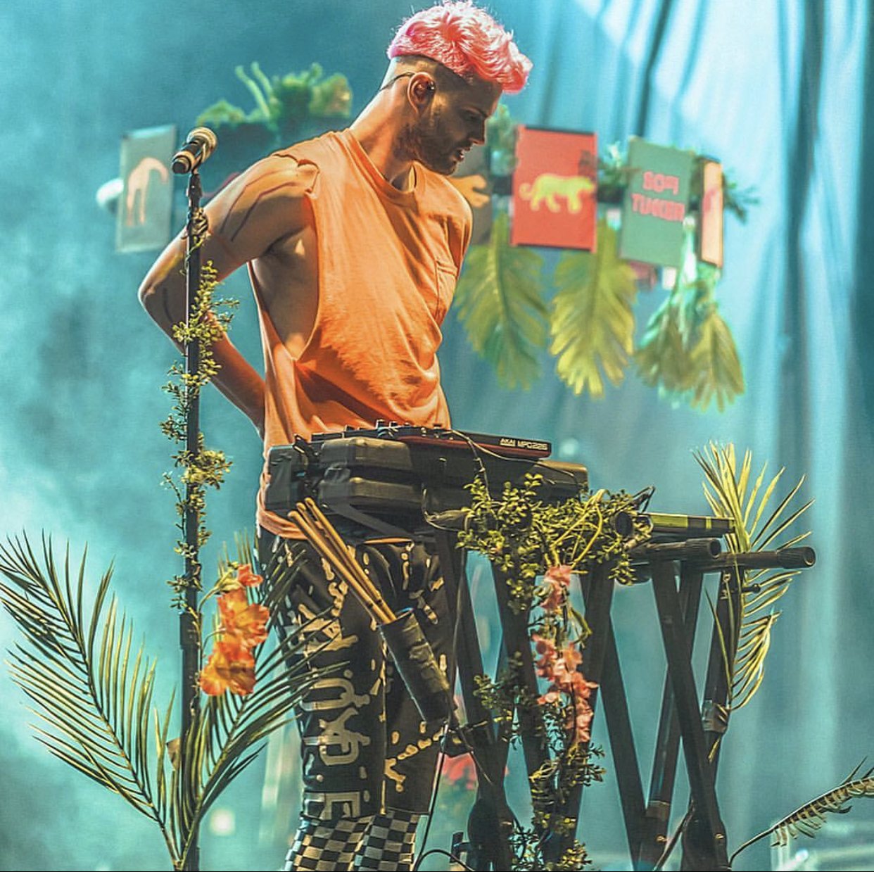 A DJ with pink hair and a sleeveless orange shirt standing behind a keyboard decorated with tropical leaves and flowers, with colorful signs and tropical leaves in the background.