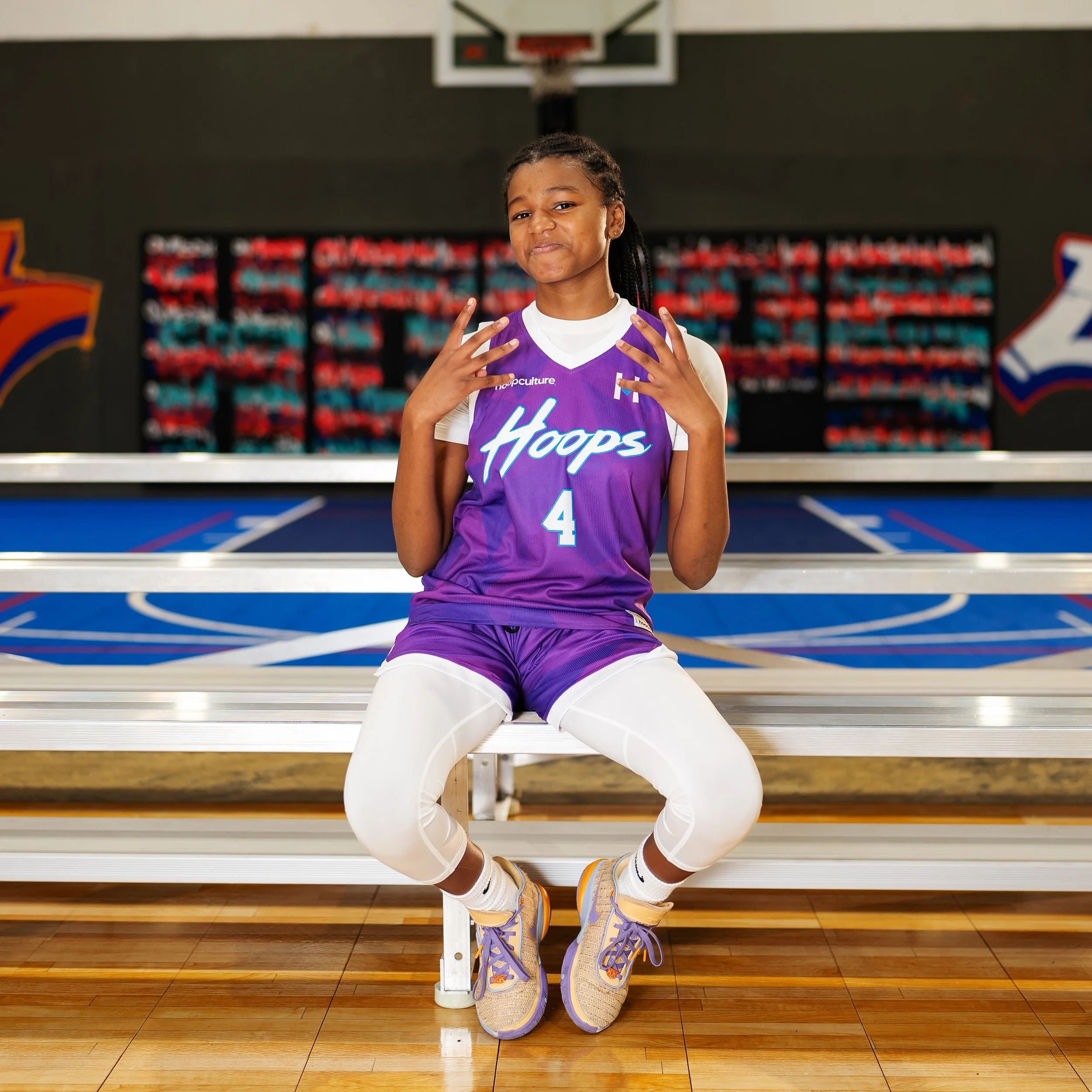 A young girl wearing a purple volleyball uniform with the number 4, sitting on a bench in a gymnasium, with her hands making a gesture and a basketball court behind her.