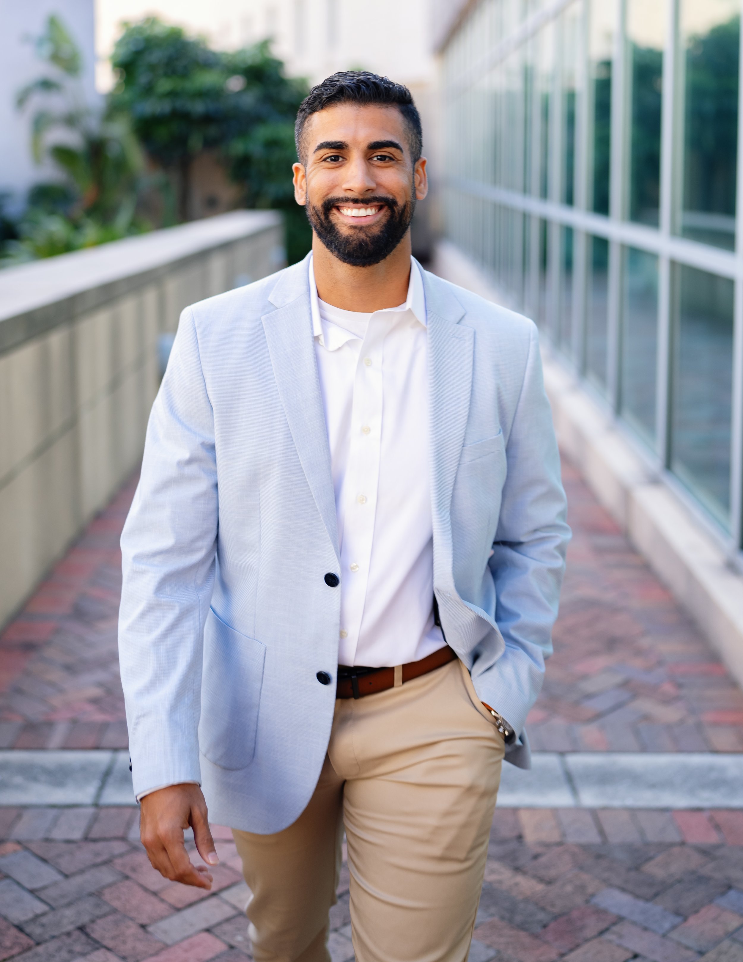 A man with a beard and short dark hair, smiling, dressed in a light blue blazer, white shirt, beige pants, standing outdoors on a brick pathway with glass windows and greenery in the background.