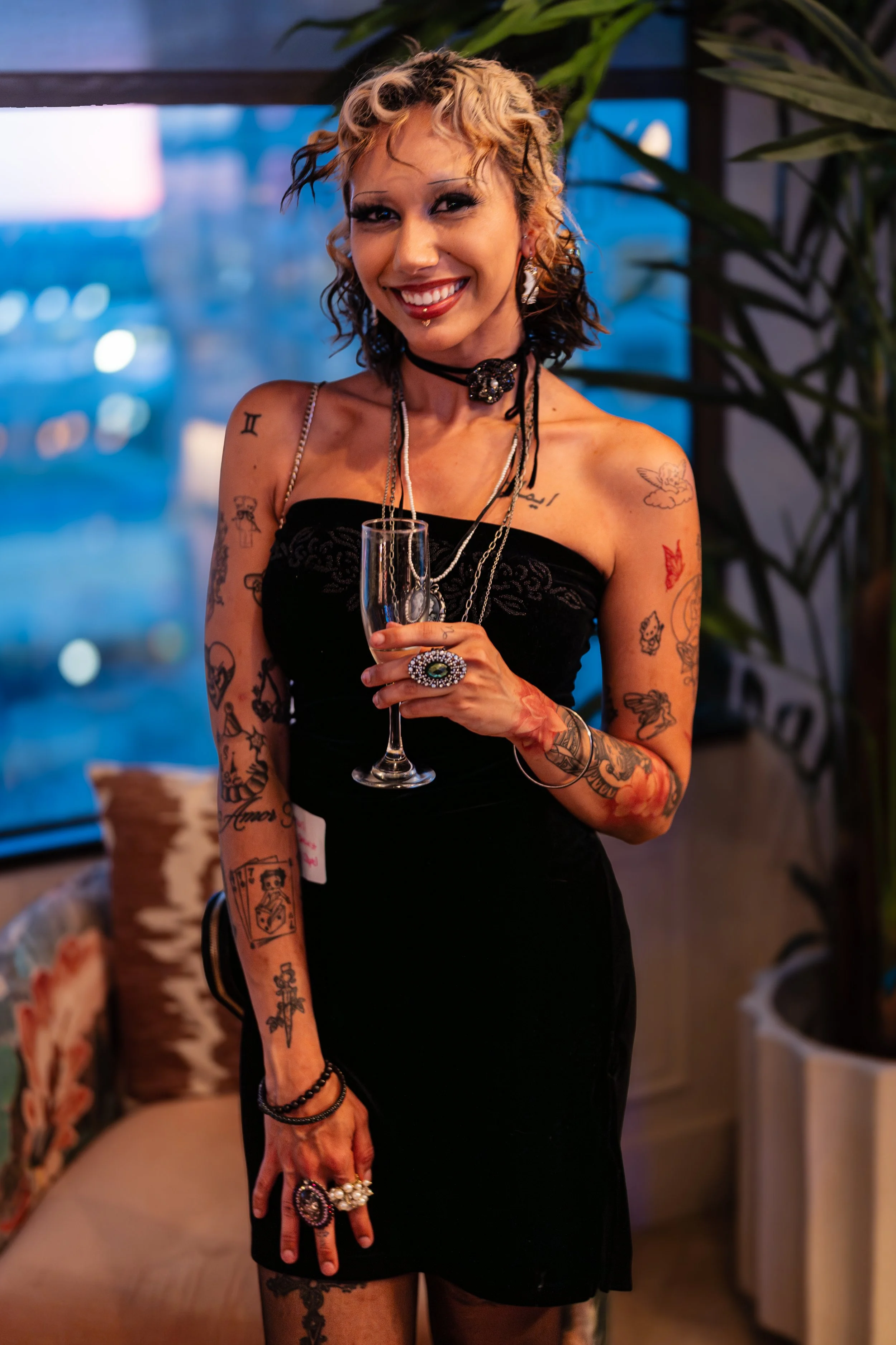 A smiling woman with tattoos on her arms and shoulders, with curly, two-toned hair, wearing a black strapless dress and multiple rings, holding a glass of champagne in an indoor setting with plants and blurred city lights in the background.
