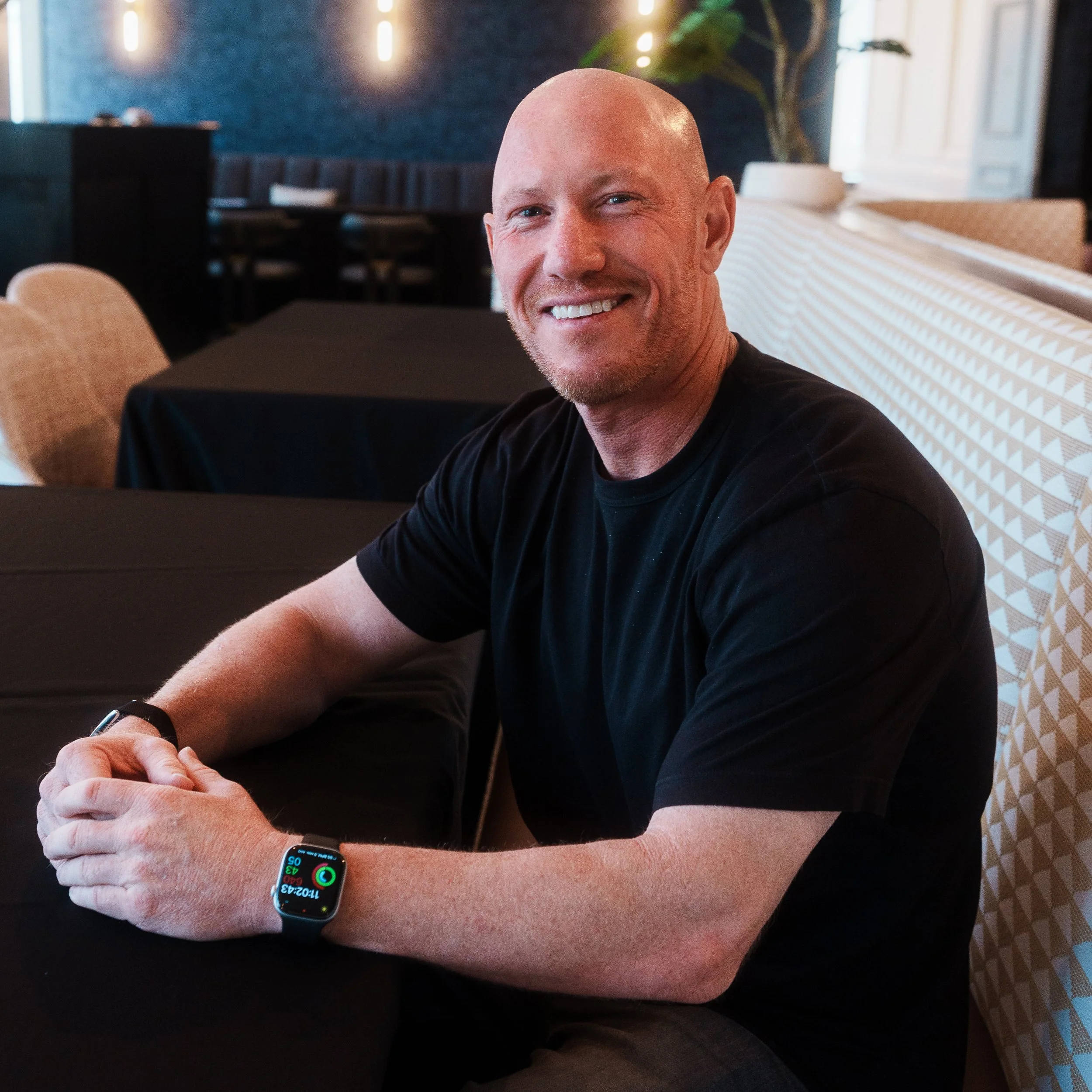 A smiling bald man with a goatee wearing a black T-shirt and a smartwatch on his left wrist, seated at a table in a modern restaurant with a blue textured wall, green plant, and ambient lighting in the background.