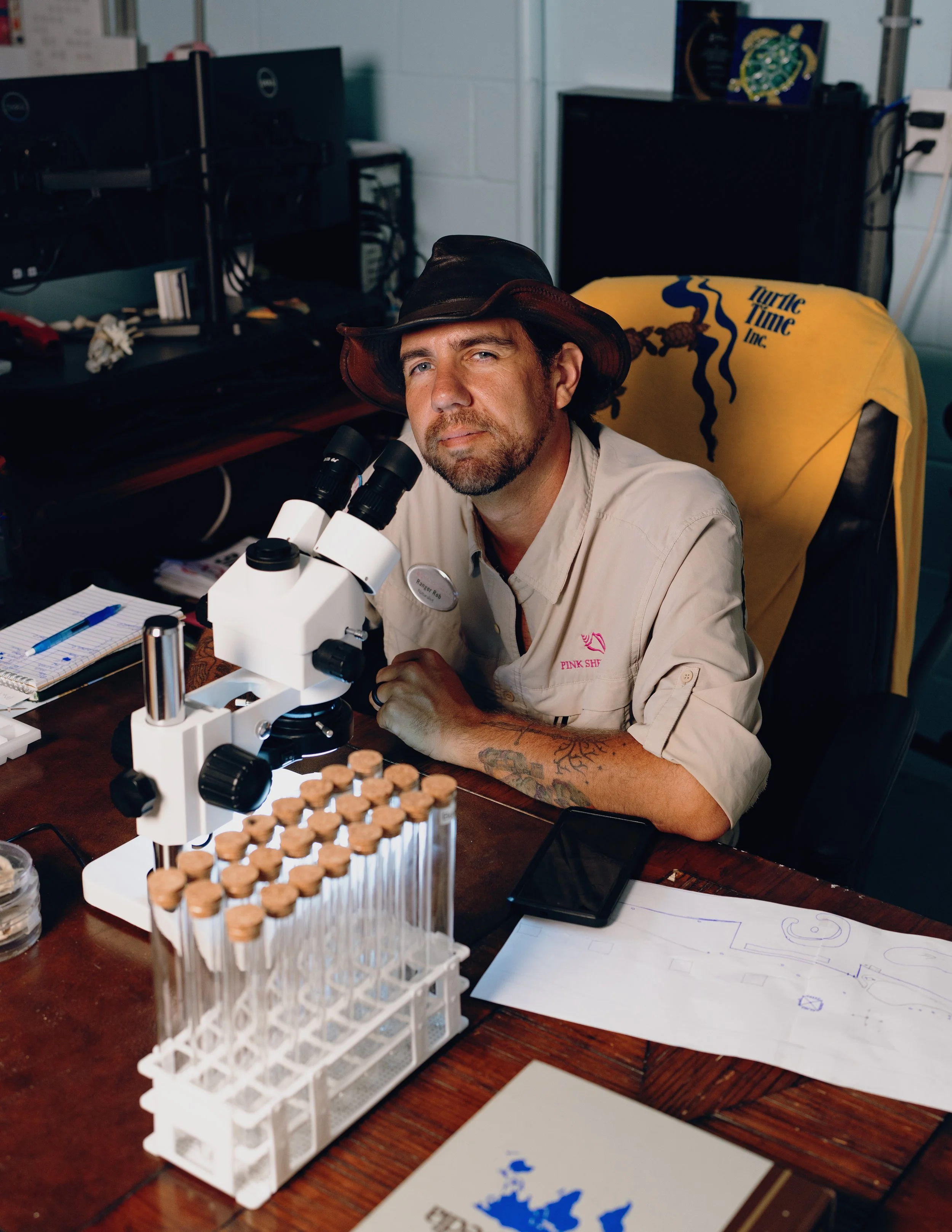 Male scientist with a tattoo on his left arm, wearing a beige shirt and a black hat, sitting at a wooden table with a microscope and test tubes, in a laboratory environment.