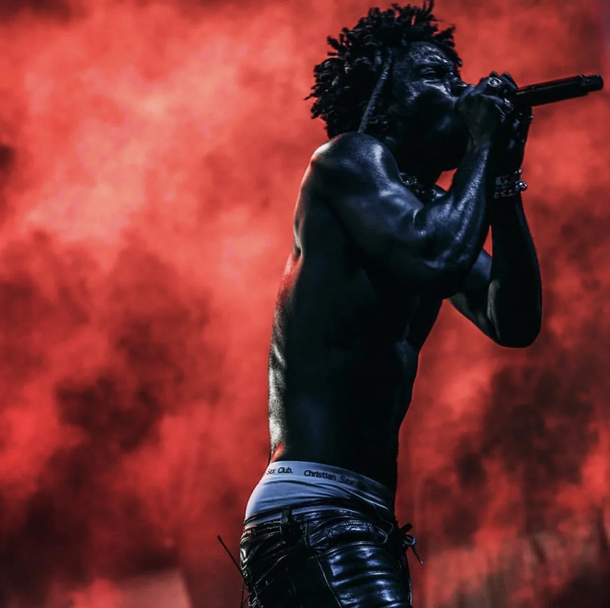 Male singer with dreadlocks passionately singing into microphone on stage with red smoky background.