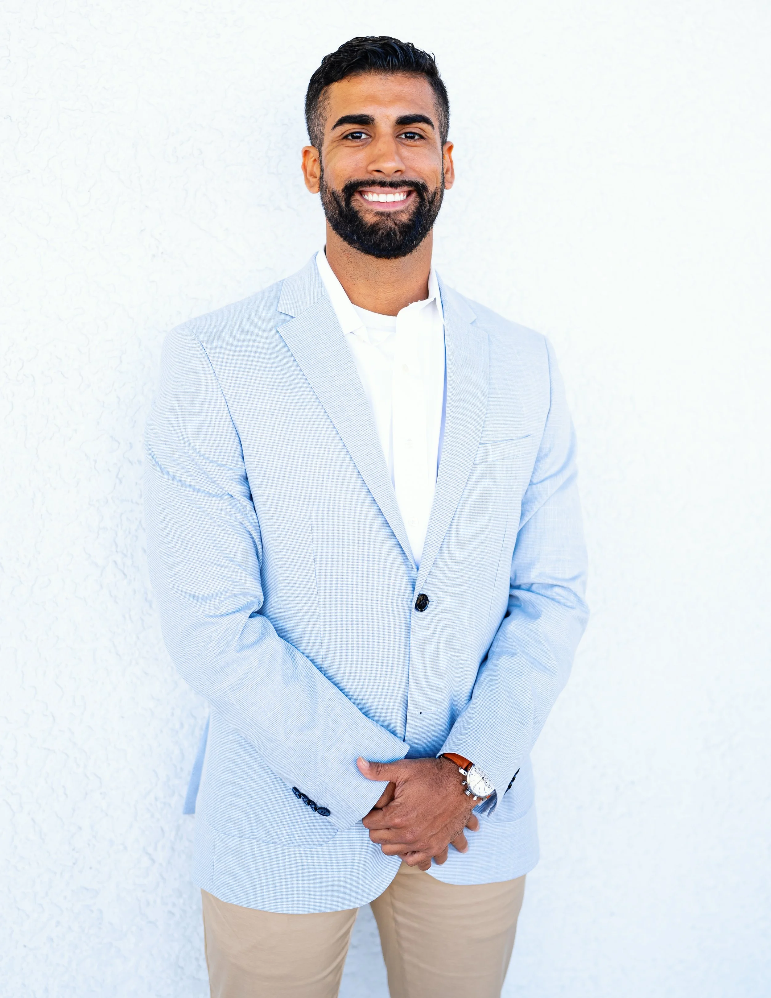 A smiling man dressed in a light blue blazer, white shirt, and beige pants standing against a plain white wall.