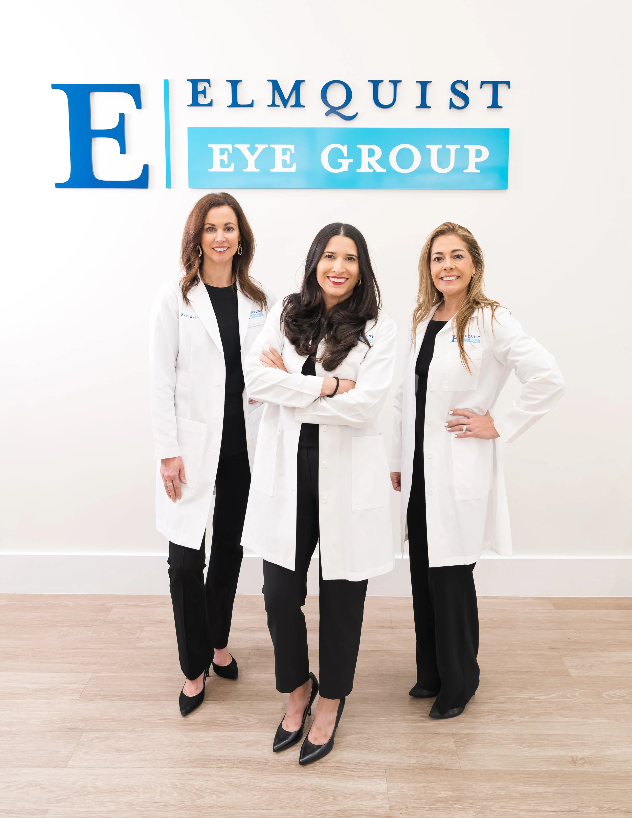 Three women wearing white lab coats standing in front of a sign that reads "Elquist Eye Group" with a white wall and wooden floor background.