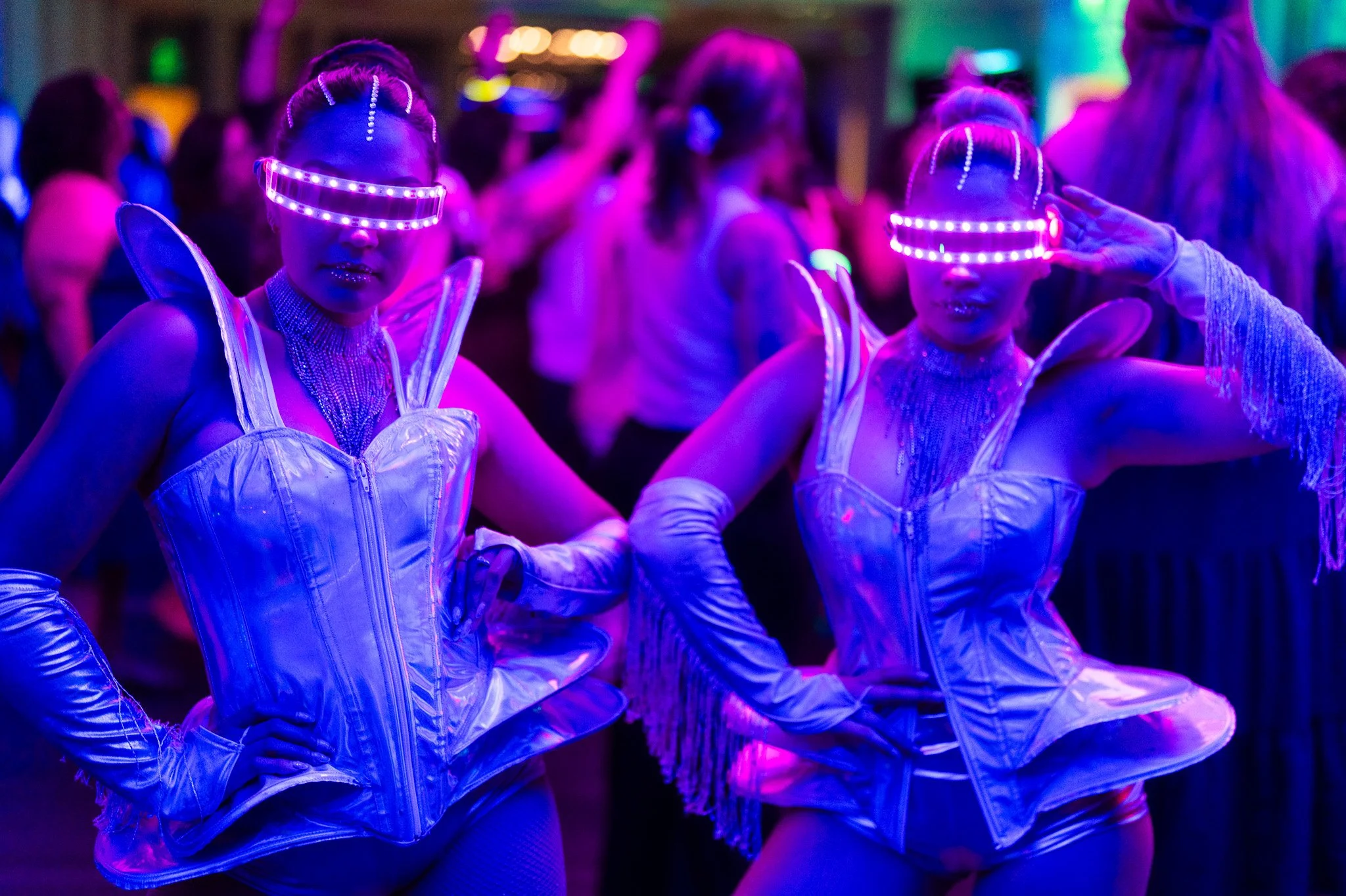 Two women dressed in futuristic costumes with illuminated visors, posing at a party with colorful neon lighting. They are wearing shiny, metallic outfits with accessories.
