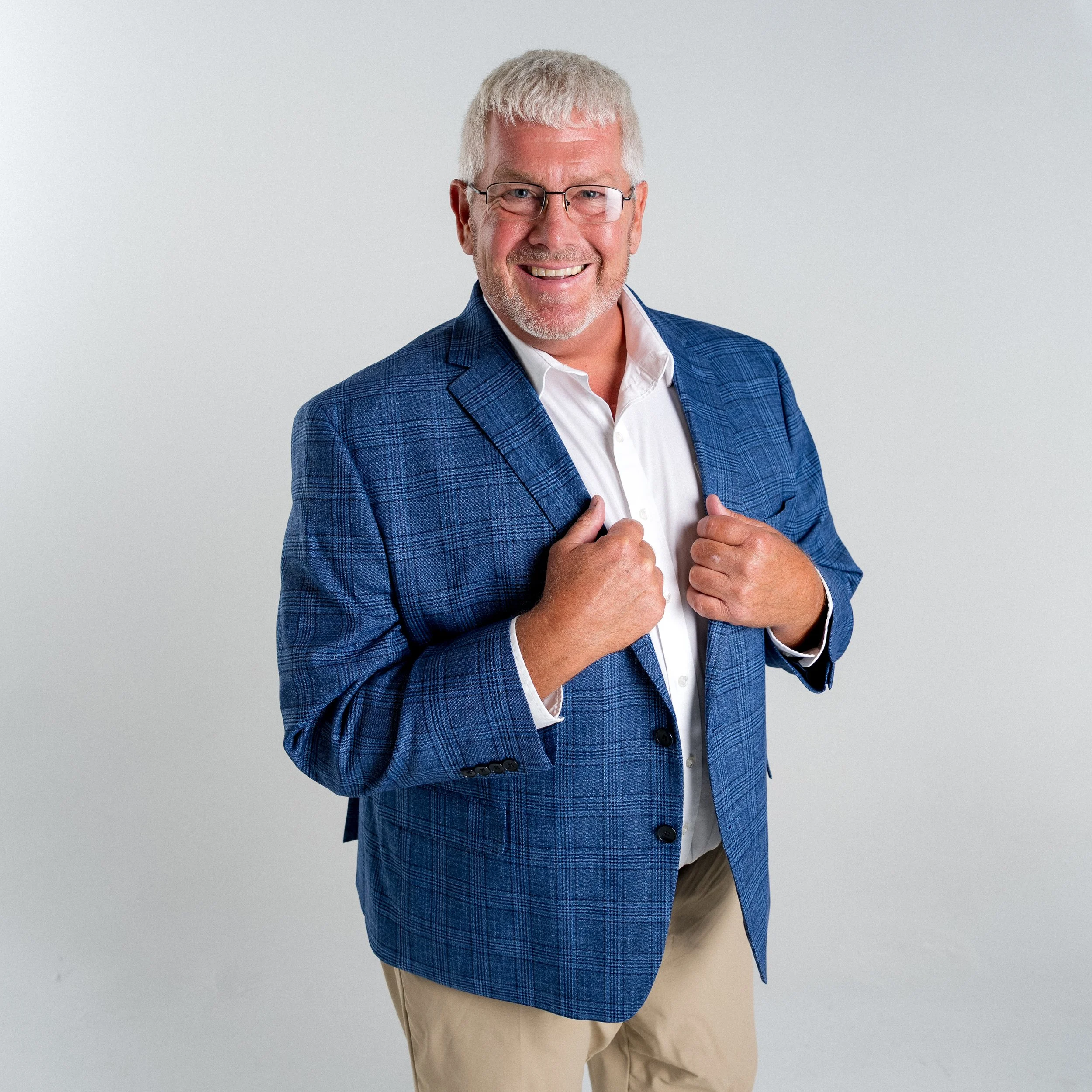 A smiling middle-aged man with gray hair, beard, and glasses, wearing a blue plaid blazer over a white shirt, standing against a light gray background.