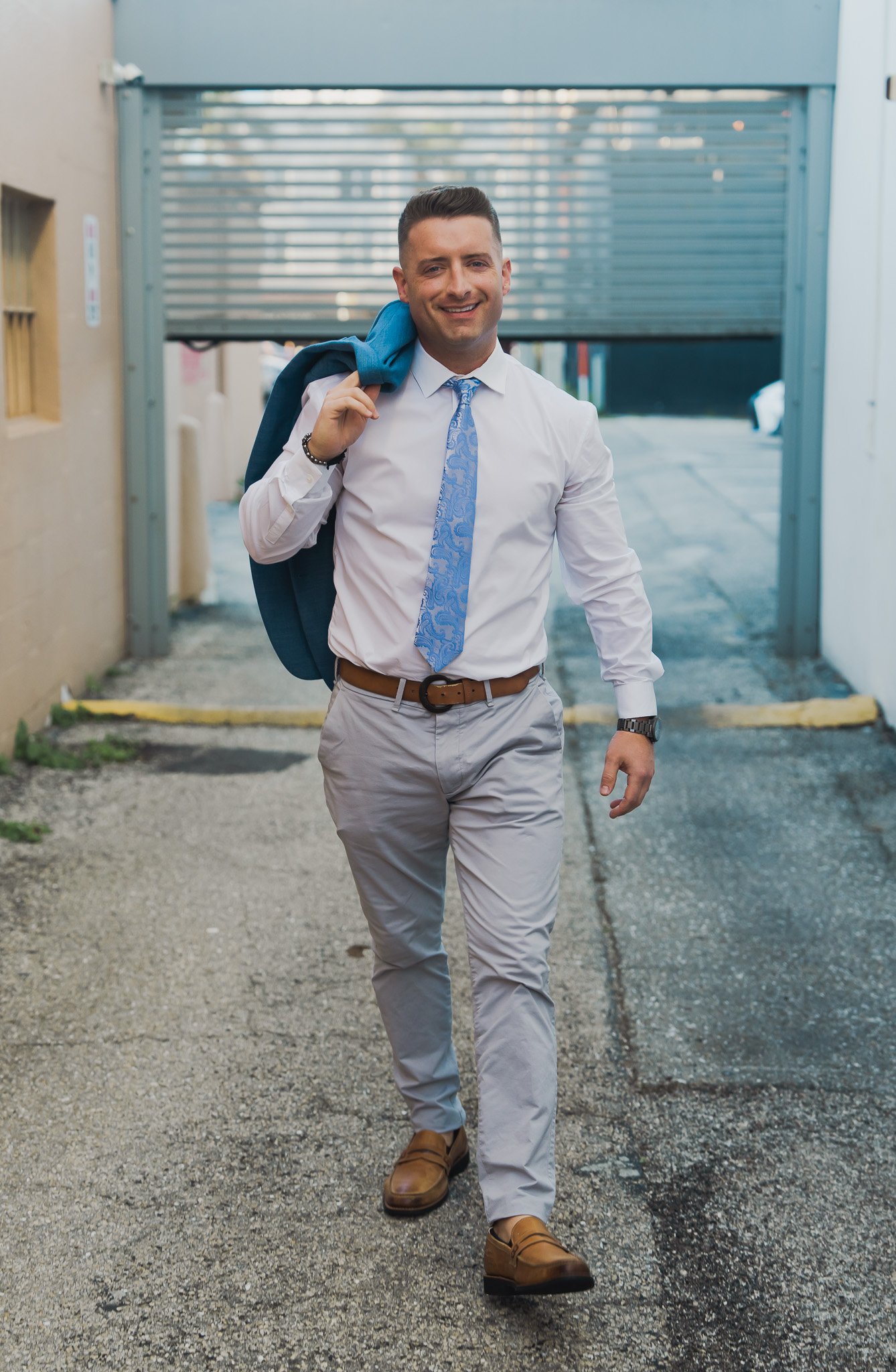 A young man in business attire walking outdoors, carrying a blue jacket over his shoulder, smiling, with a parking garage in the background.