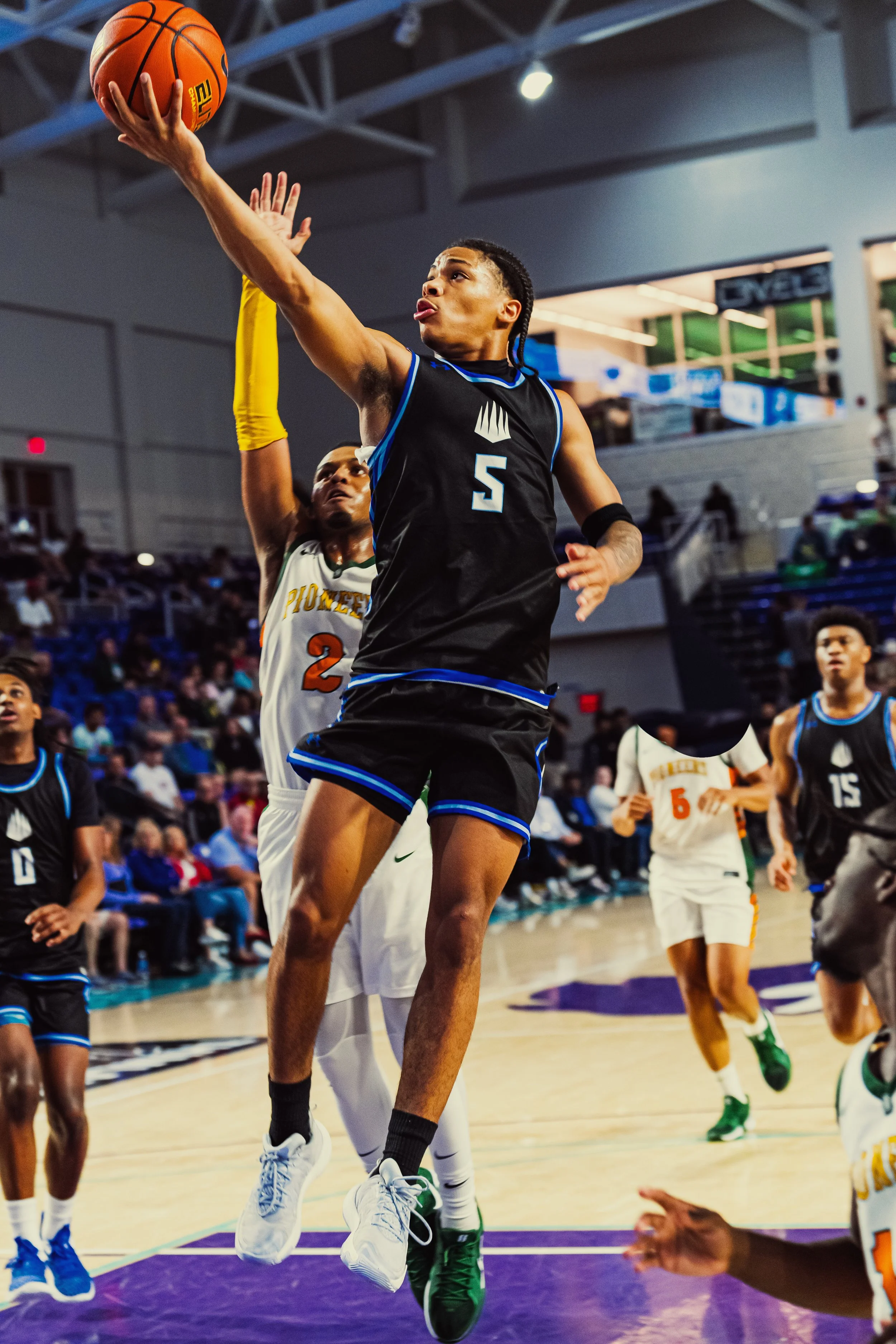 A basketball player in a black and blue uniform jumps to score while an opponent in a white jersey attempts to block the shot during a game in an indoor arena.