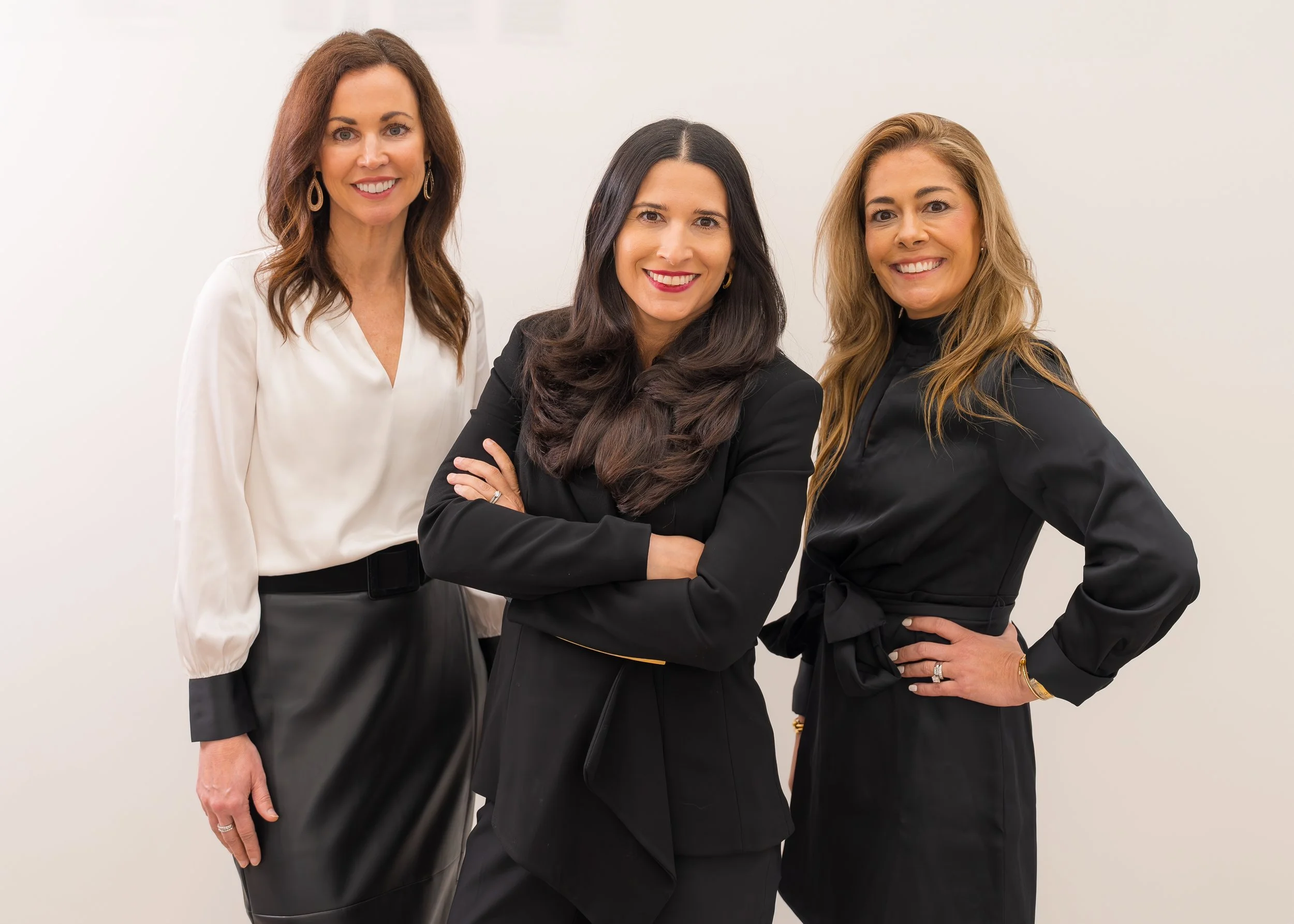 Three women standing together against a plain white wall, smiling and looking at the camera. They are dressed in professional attire with dark and white blouses and skirts or pants.