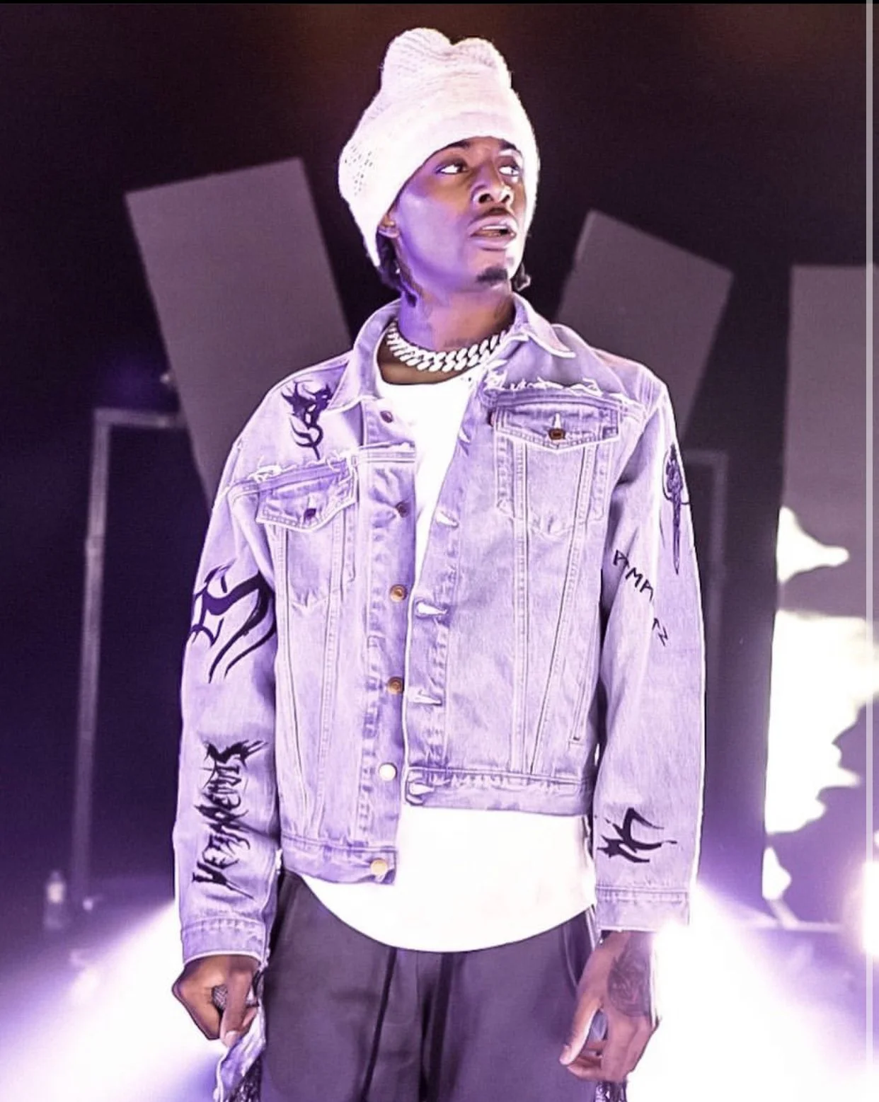 A young man with a white beanie, gold chain necklace, and a purple jacket with black graffiti-style writing, standing in a studio with bright lights and dark background.