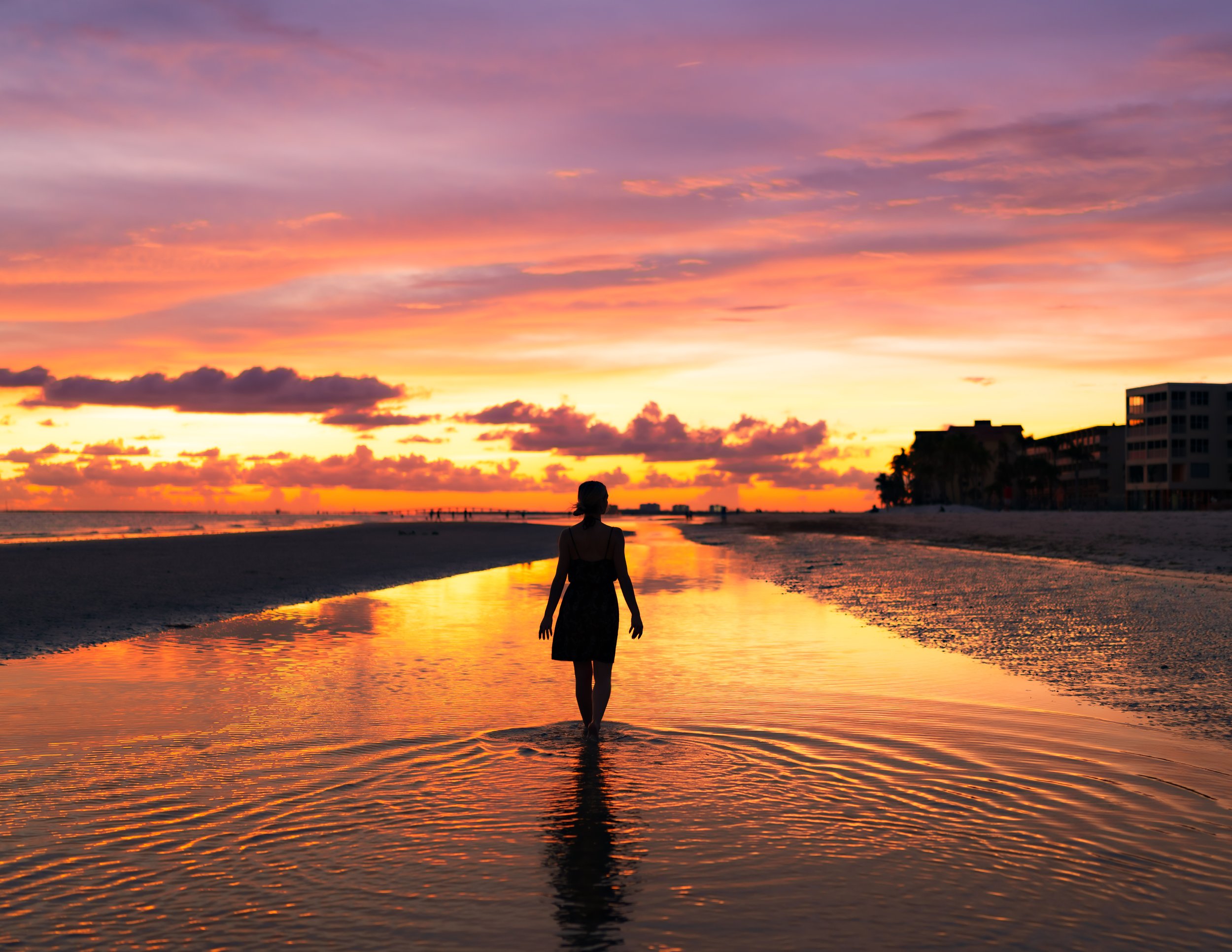A person walking along a beach at sunset with colorful sky and clouds reflected in shallow water.
