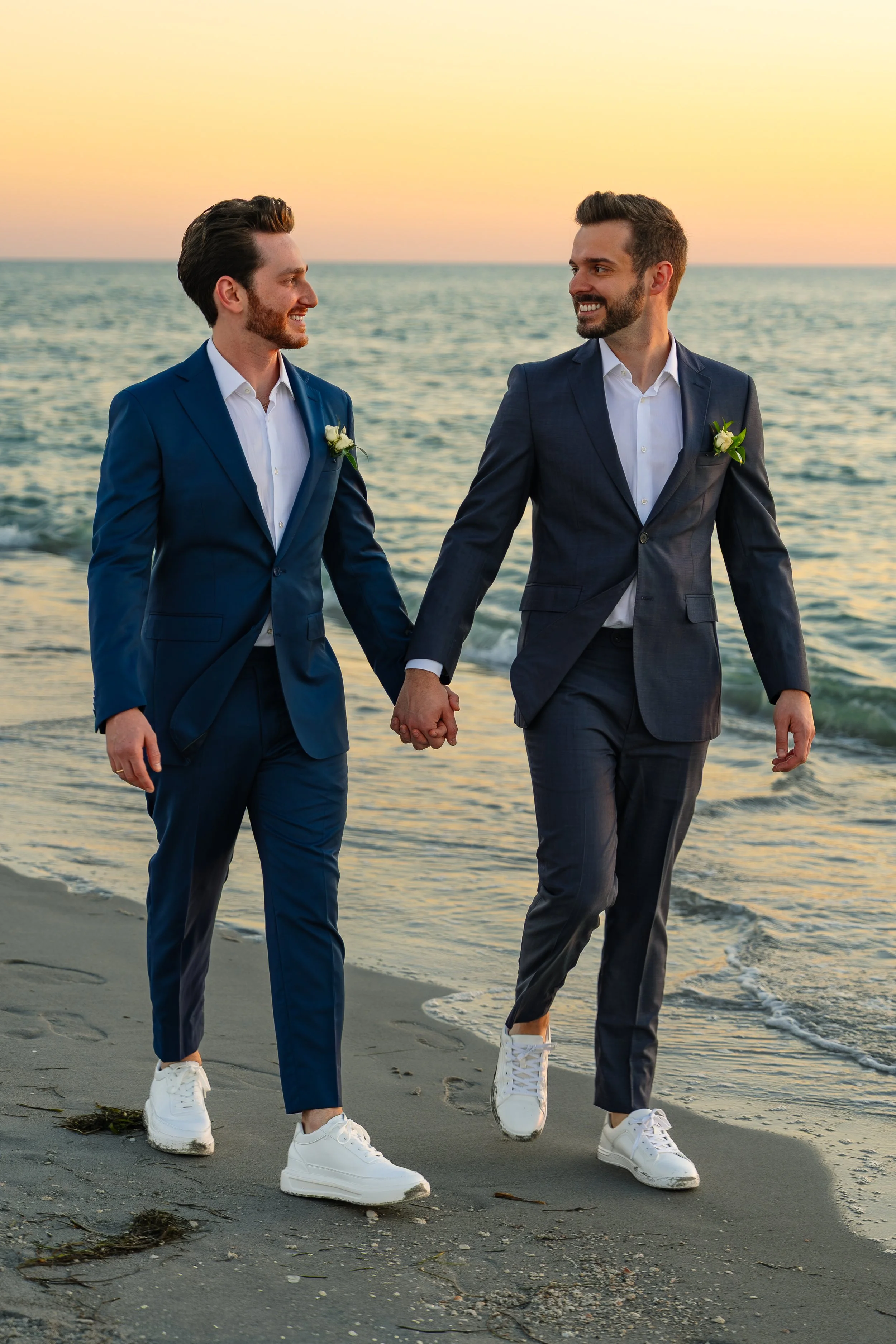 Two men in suits holding hands and walking on a beach during sunset, smiling at each other.