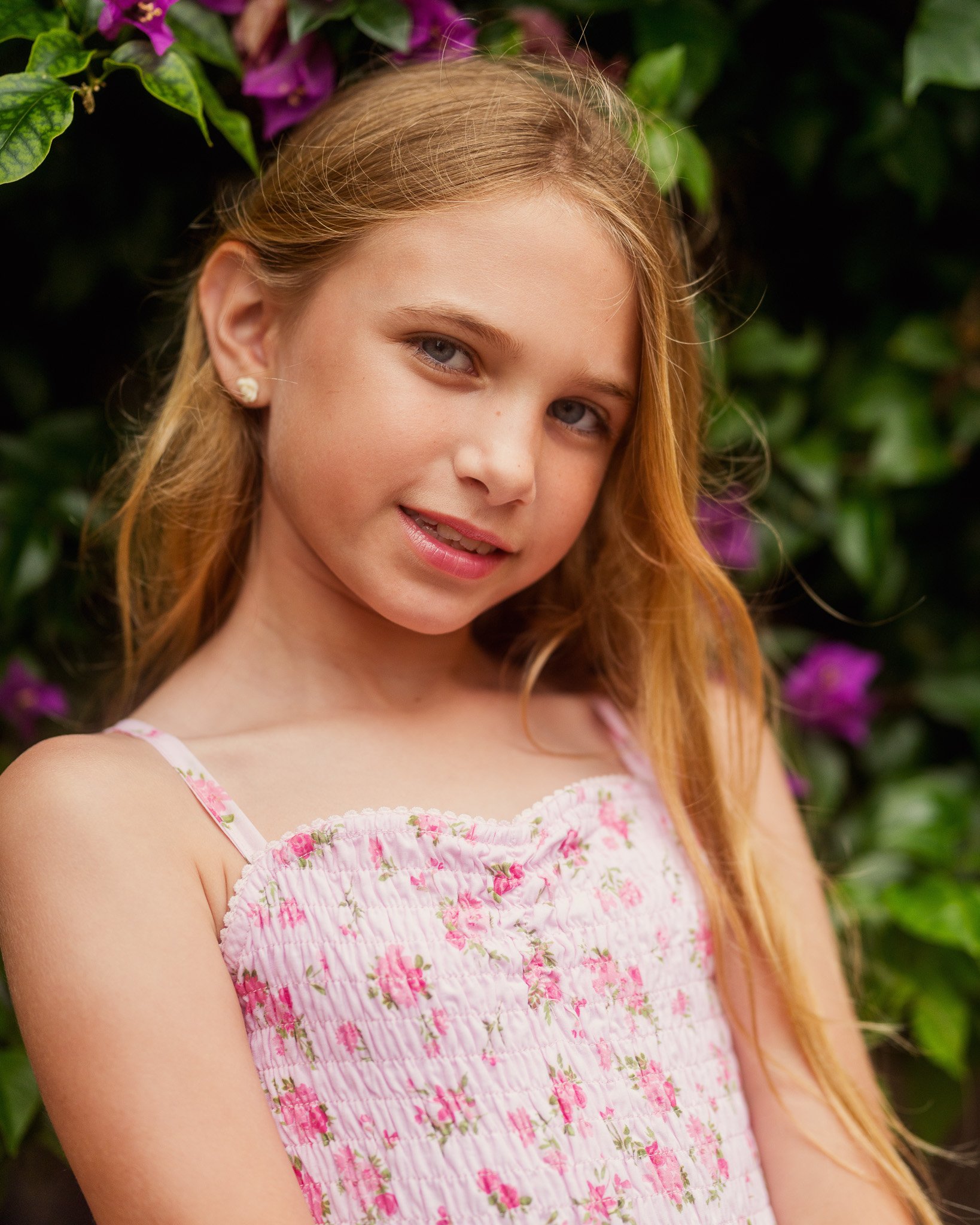 Close-up of a young girl with long strawberry blonde hair, wearing a pink floral dress, standing in front of green foliage and purple flowers.