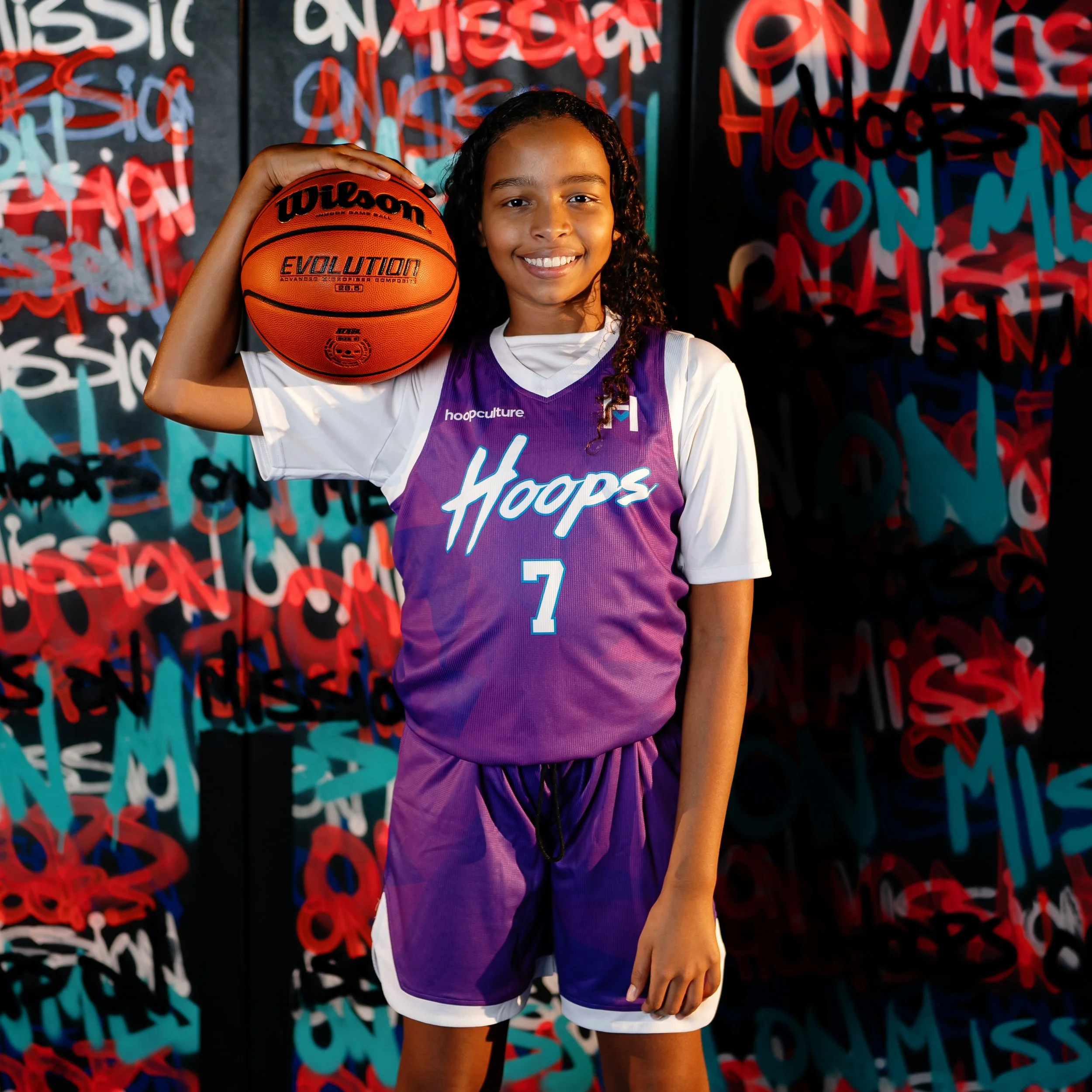 A young girl smiling while holding a basketball in front of a colorful graffiti background.