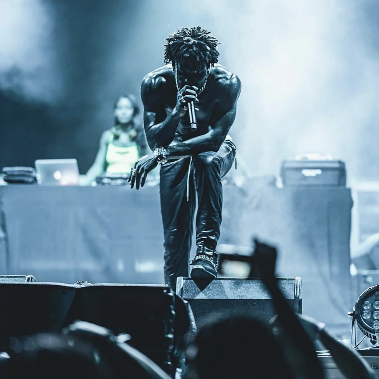 A shirtless male performer with dreadlocks holding a microphone and raising one foot on stage in a concert setting with blue lighting.