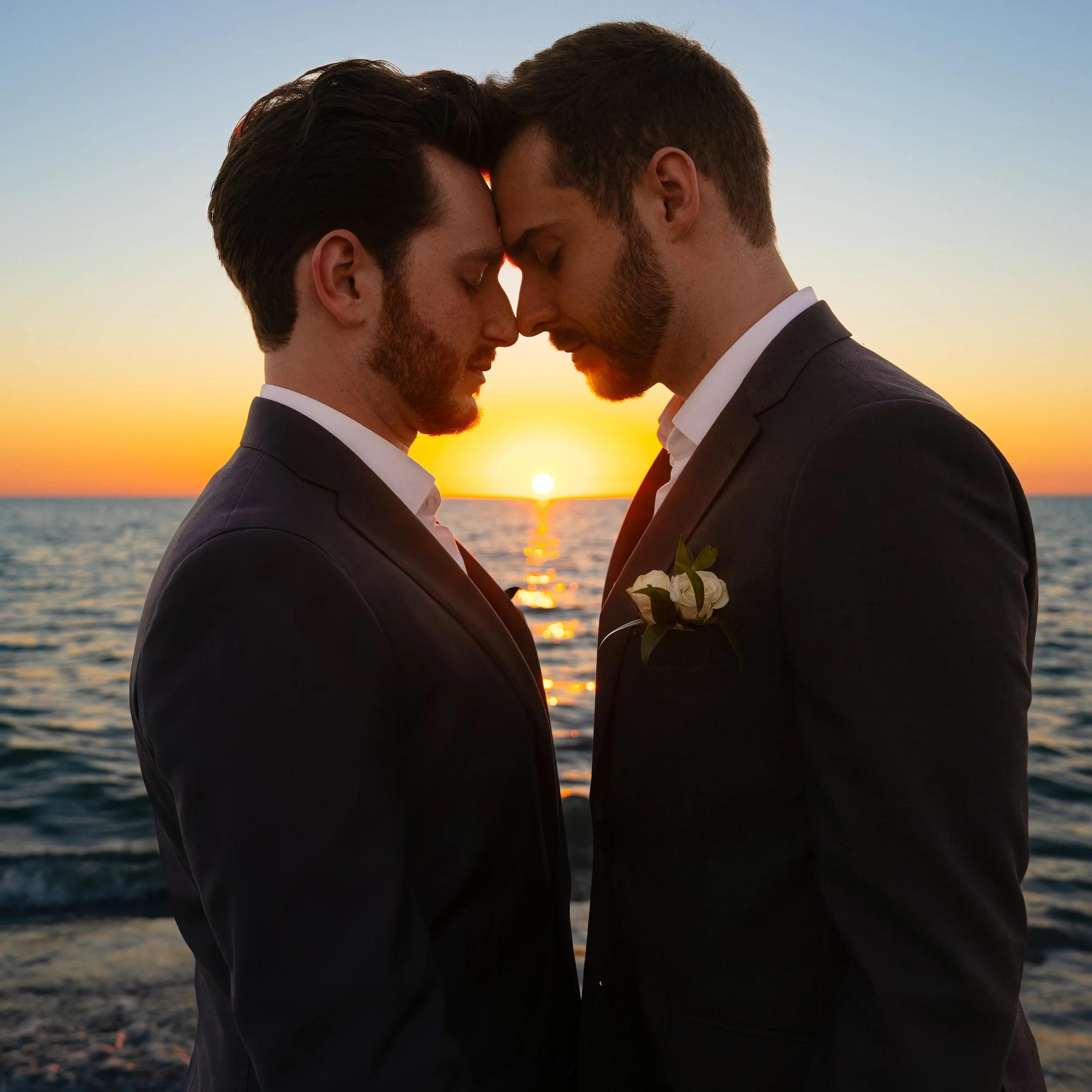 Two men in suits standing close with foreheads touching during a sunset on the beach.