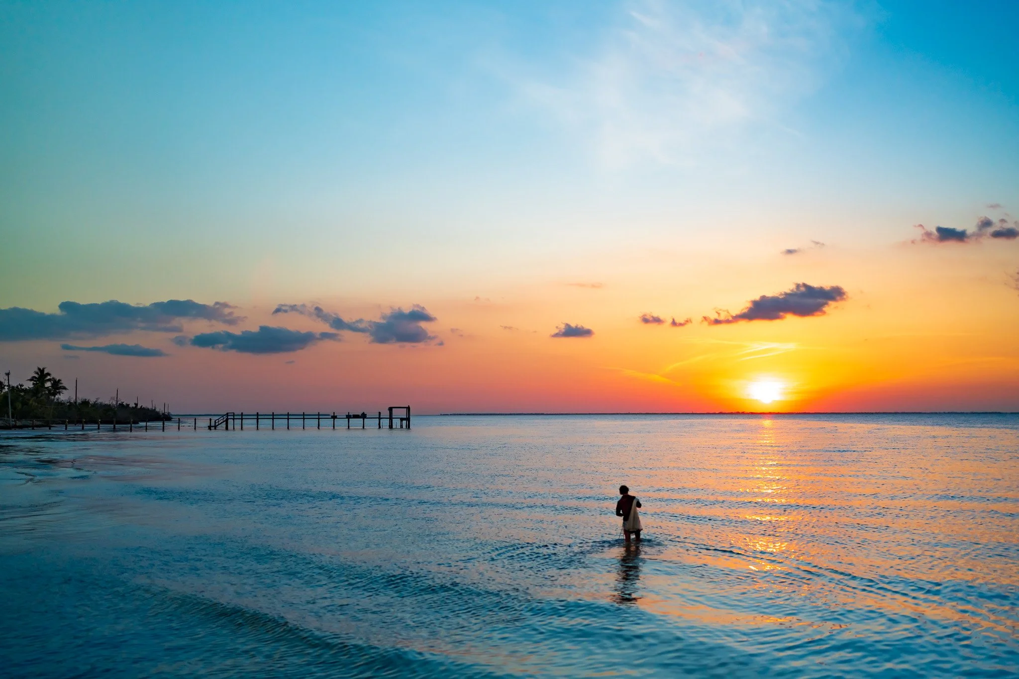 A person wading in the ocean at sunset, with a pier extending into the water and a colorful sky with clouds and a setting sun.