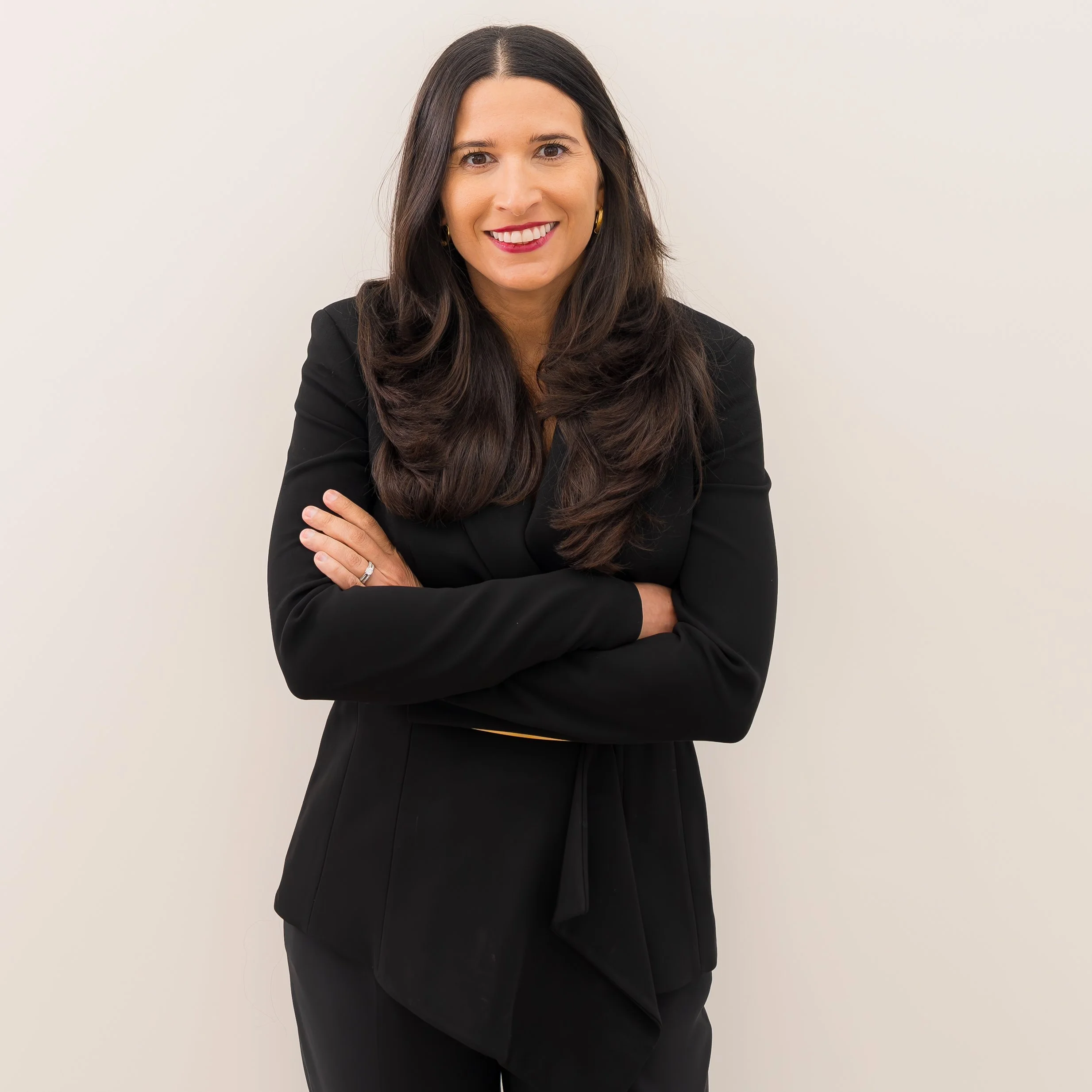 A woman with long dark hair, wearing a black blazer and smiling with arms crossed, standing against a plain white background.