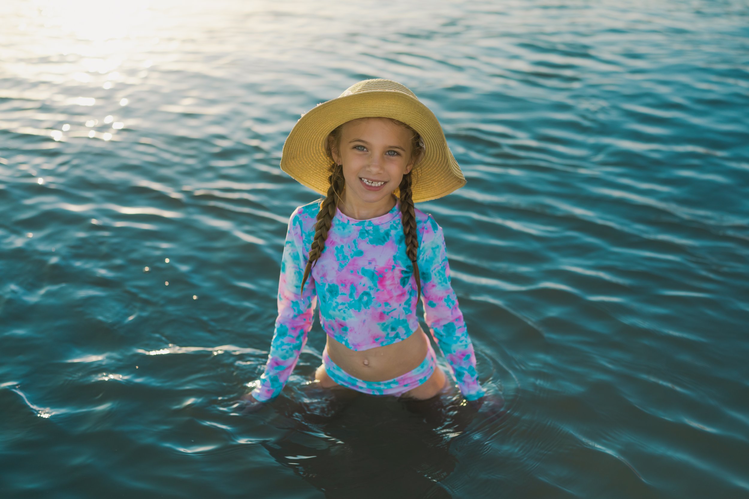 A young girl with braided hair wearing a wide-brimmed straw hat, a colorful long-sleeve swim top, and swim bottoms, standing in the water with a bright smile.