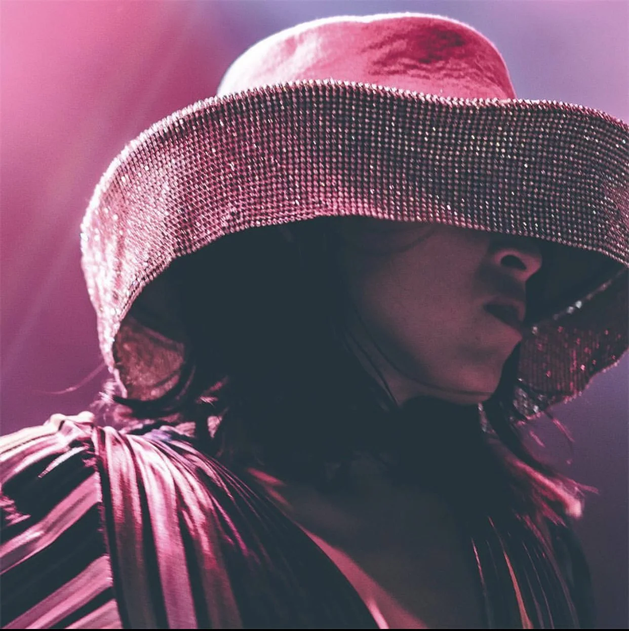 A woman wearing a large, sparkly pink hat partially covering her face, with a dark background.