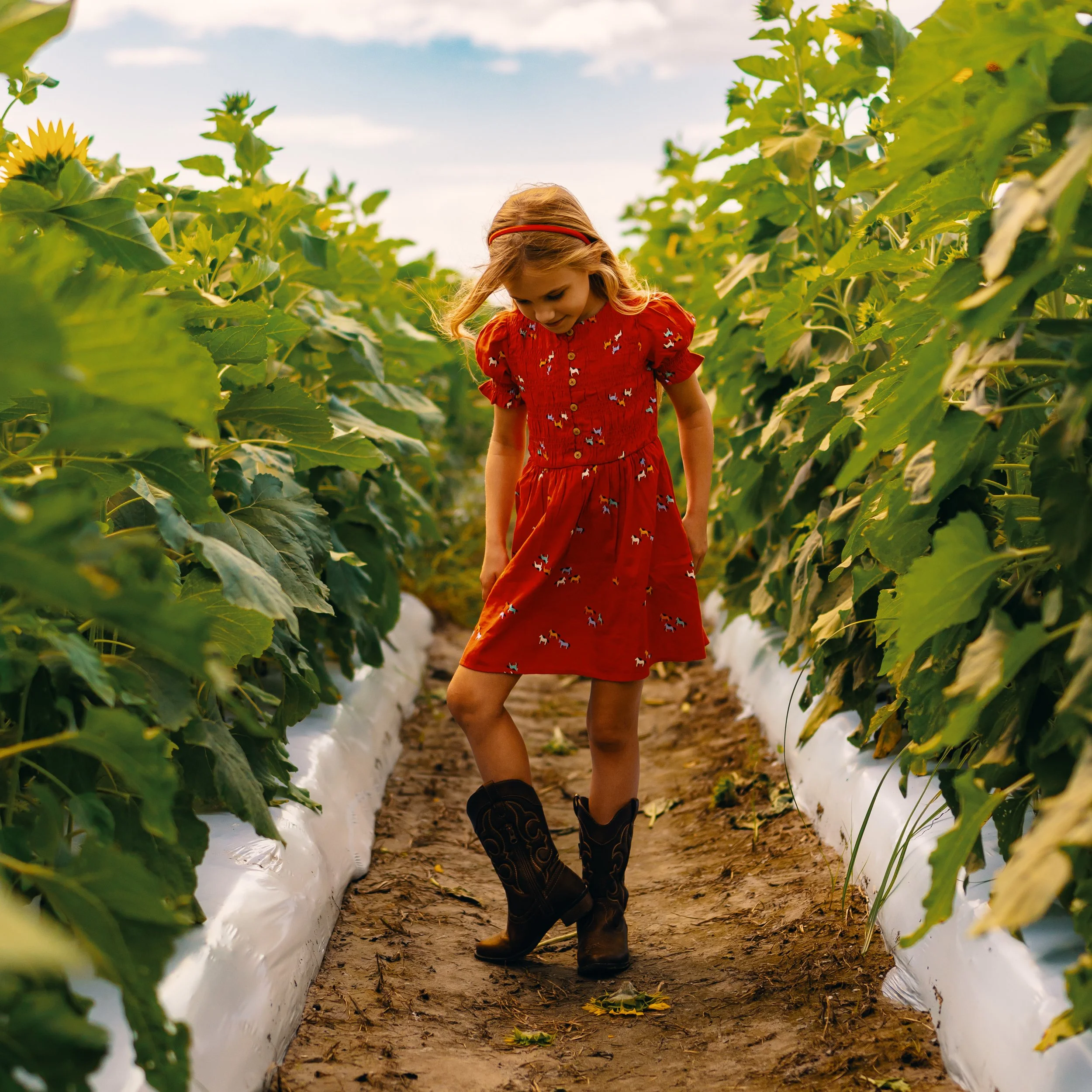A young girl in a red dress with small colorful animal patterns and black cowboy boots standing on a dirt path between rows of green plants, with a partly cloudy sky in the background.