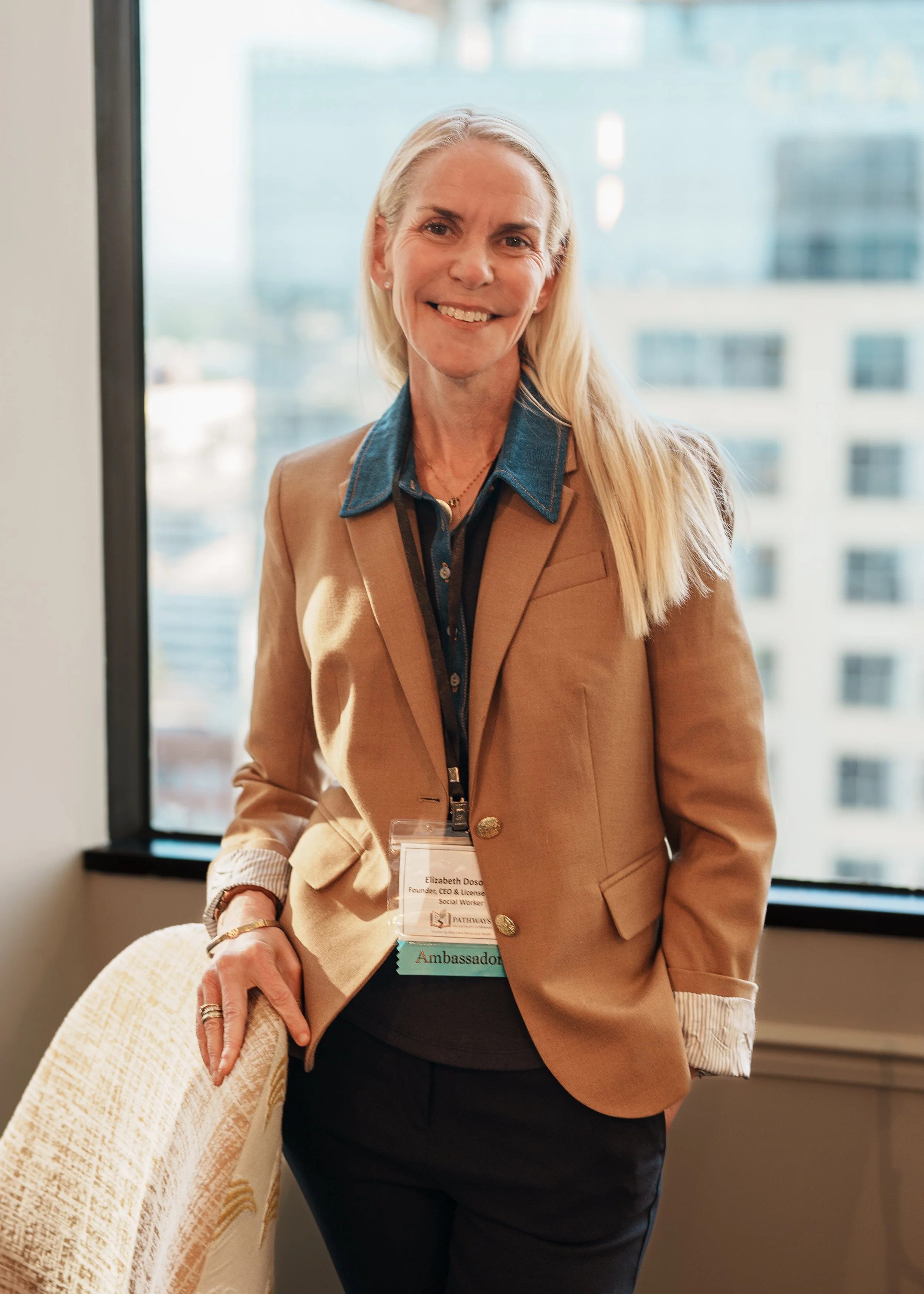 A woman with long blonde hair wearing a tan blazer and a blue shirt with a name badge, standing indoors near a window with a cityscape in the background, smiling at the camera.
