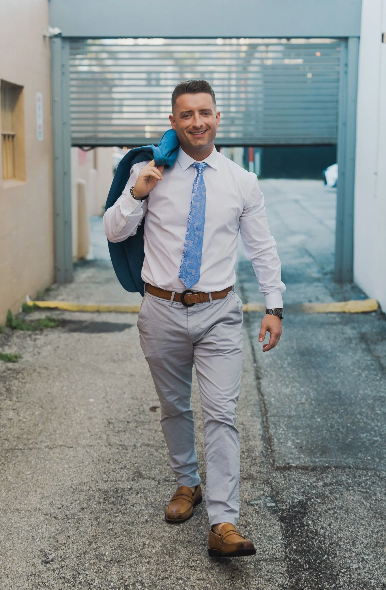 A young man in business attire walking outdoors, carrying a blue jacket over his shoulder, smiling, with a parking garage in the background.