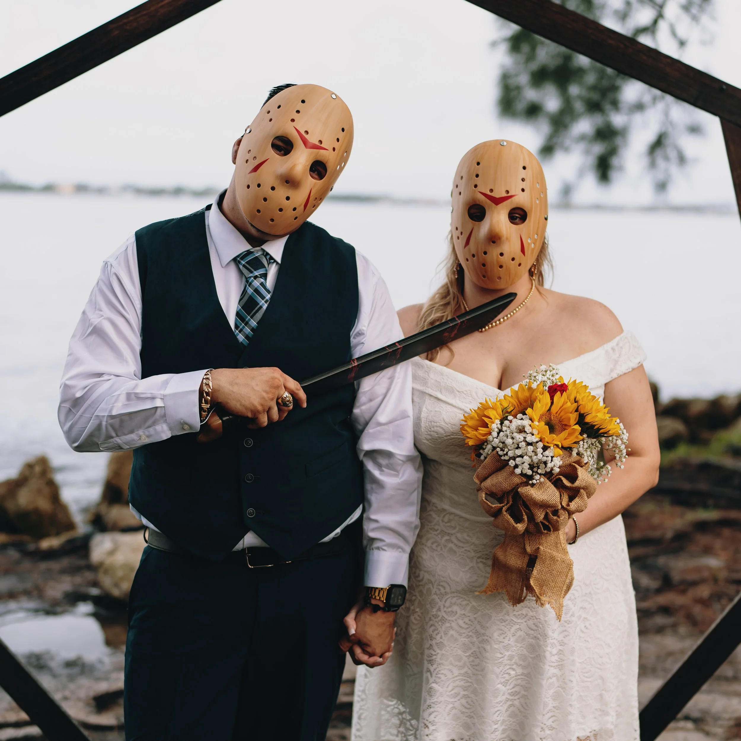 A couple dressed in wedding attire wearing horror movie Jason Voorhees masks, holding hands, standing outdoors near a body of water, during daytime. The woman is holding a bouquet of sunflowers and baby's breath.