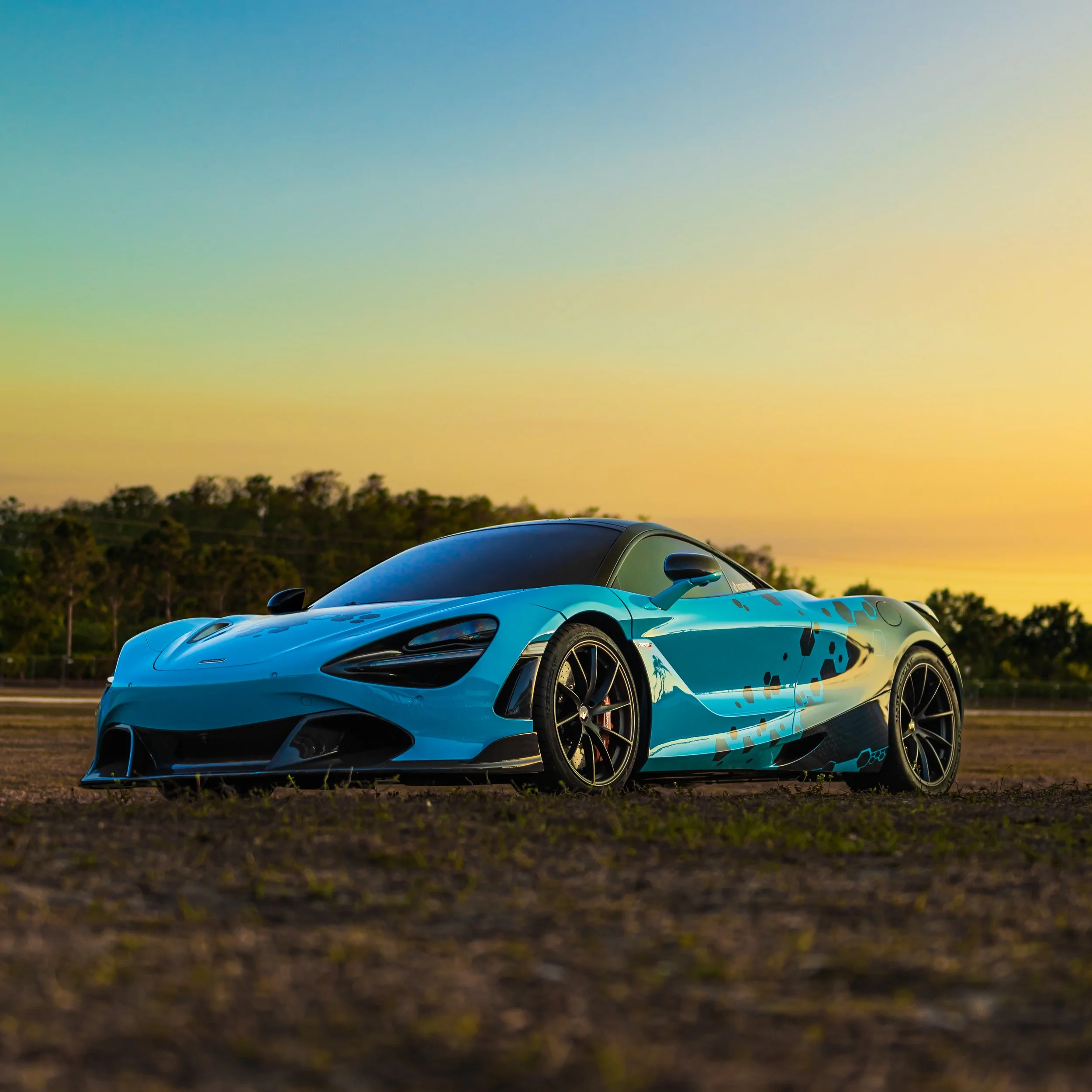 A sleek blue sports car parked on dry ground during sunset with trees in the background.