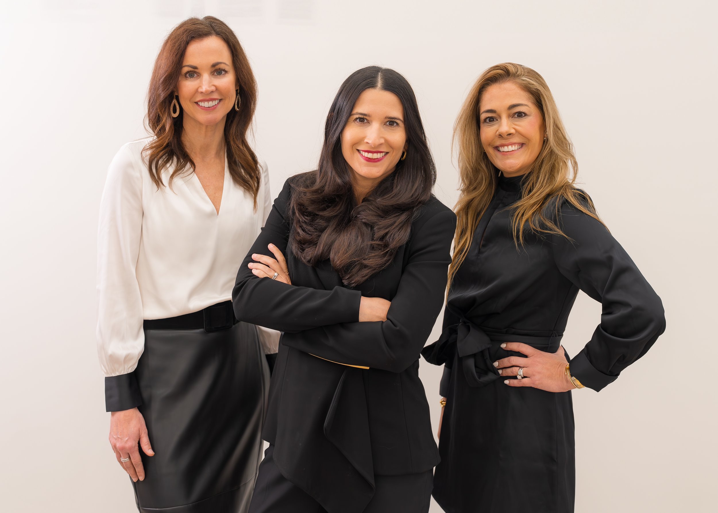 Three women standing together against a plain white wall, smiling and looking at the camera. They are dressed in professional attire with dark and white blouses and skirts or pants.
