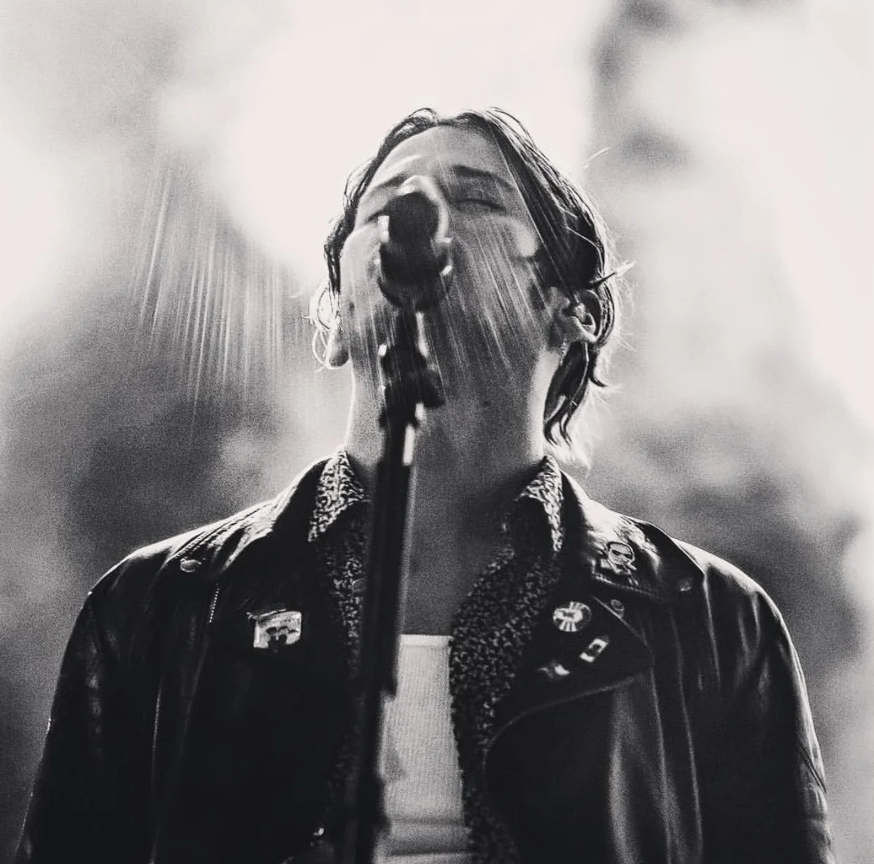 Black and white photo of a person singing into a microphone, with rain-like light streaks around them, wearing a leather jacket with pins and patches.