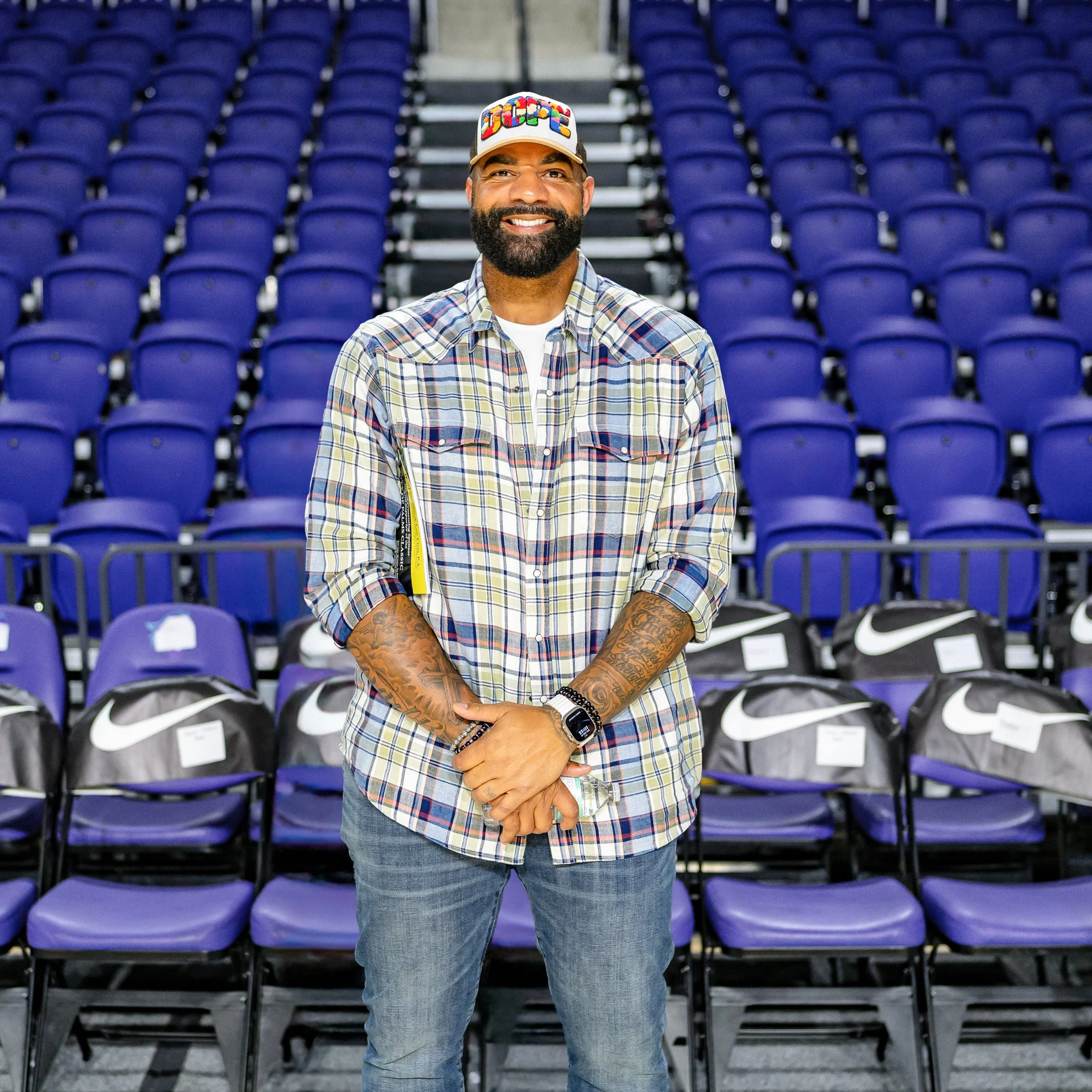 A man with a beard and tattoos stands in an arena with rows of empty blue seats behind him, wearing a plaid shirt, a colorful cap, and smiling.