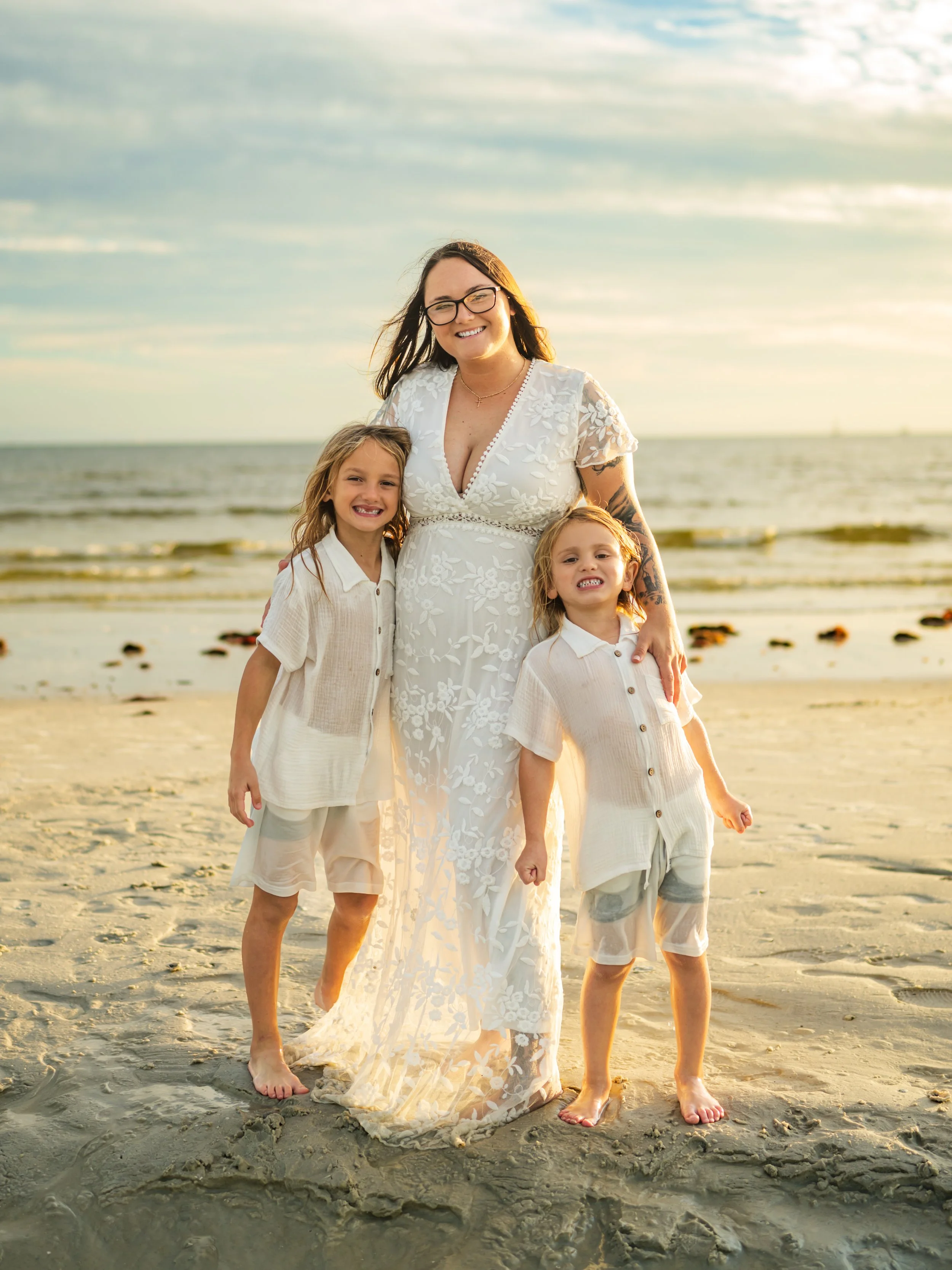 A woman in a white lace dress with glasses standing on the beach with two young girls in light-colored outfits.