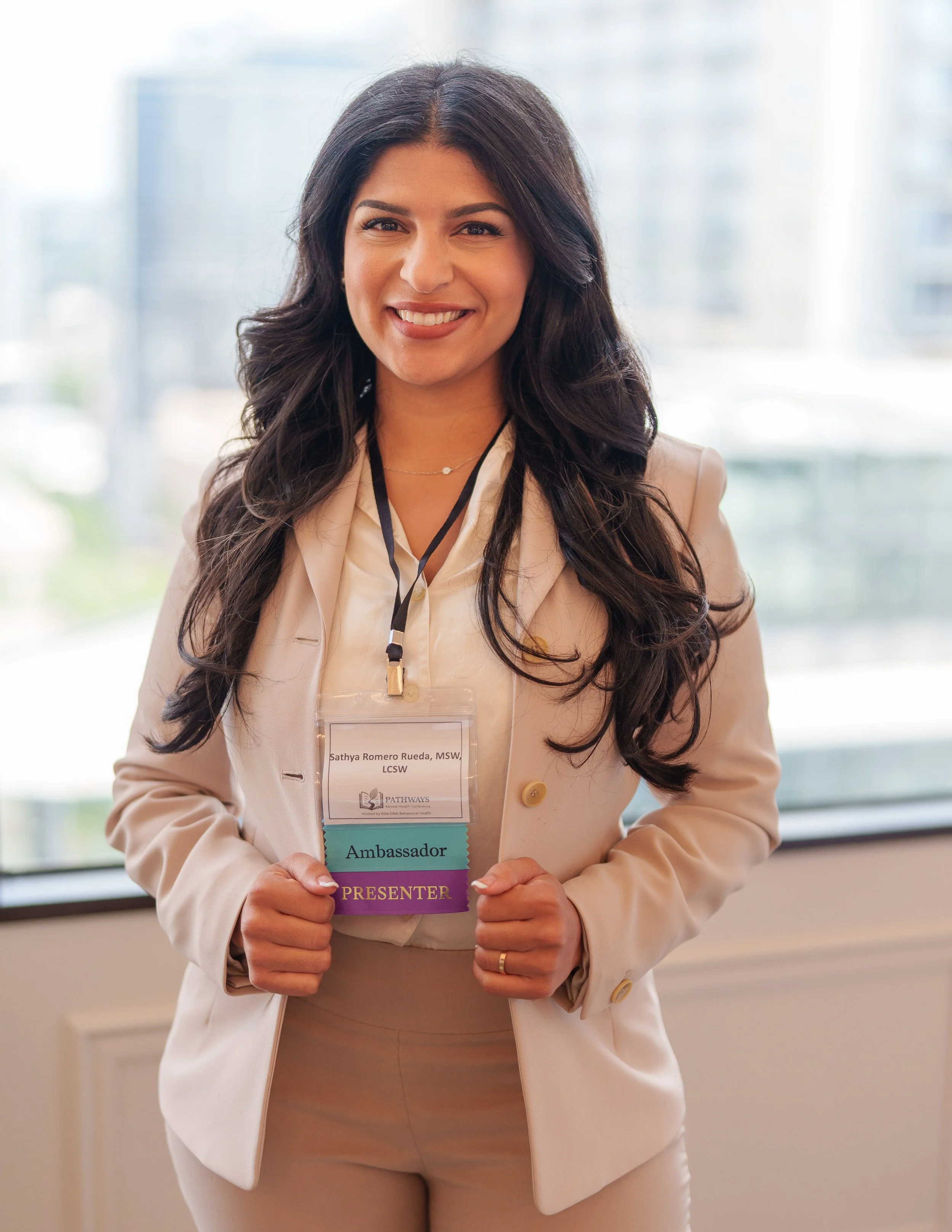 A woman in professional attire holding a conference badge that reads 'Ambassador' and 'Presenter', smiling, with a blurred cityscape in the background.