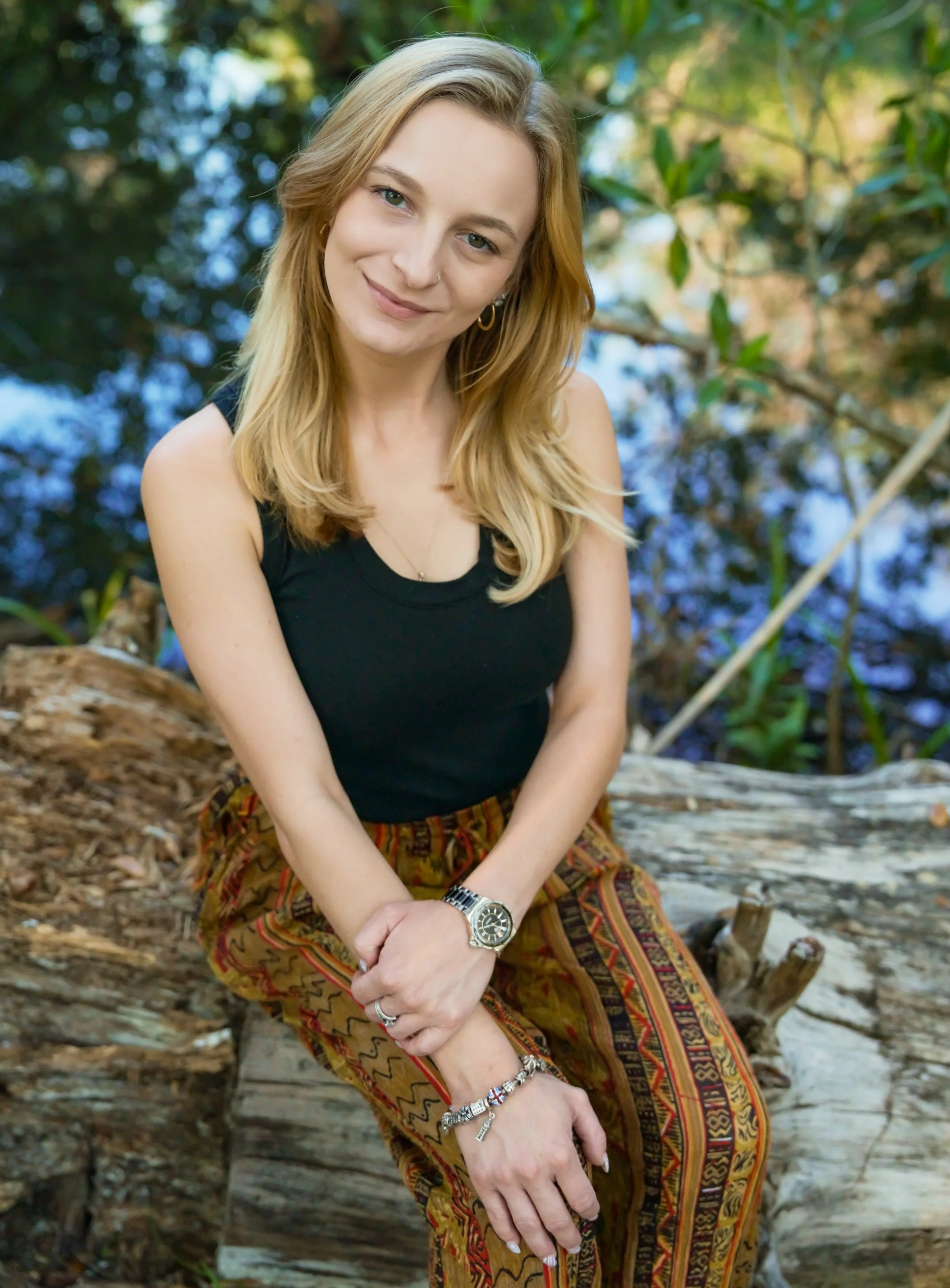 A young woman with blonde hair, wearing a black tank top and patterned pants, sitting on a fallen log in a wooded area, smiling and looking at the camera.