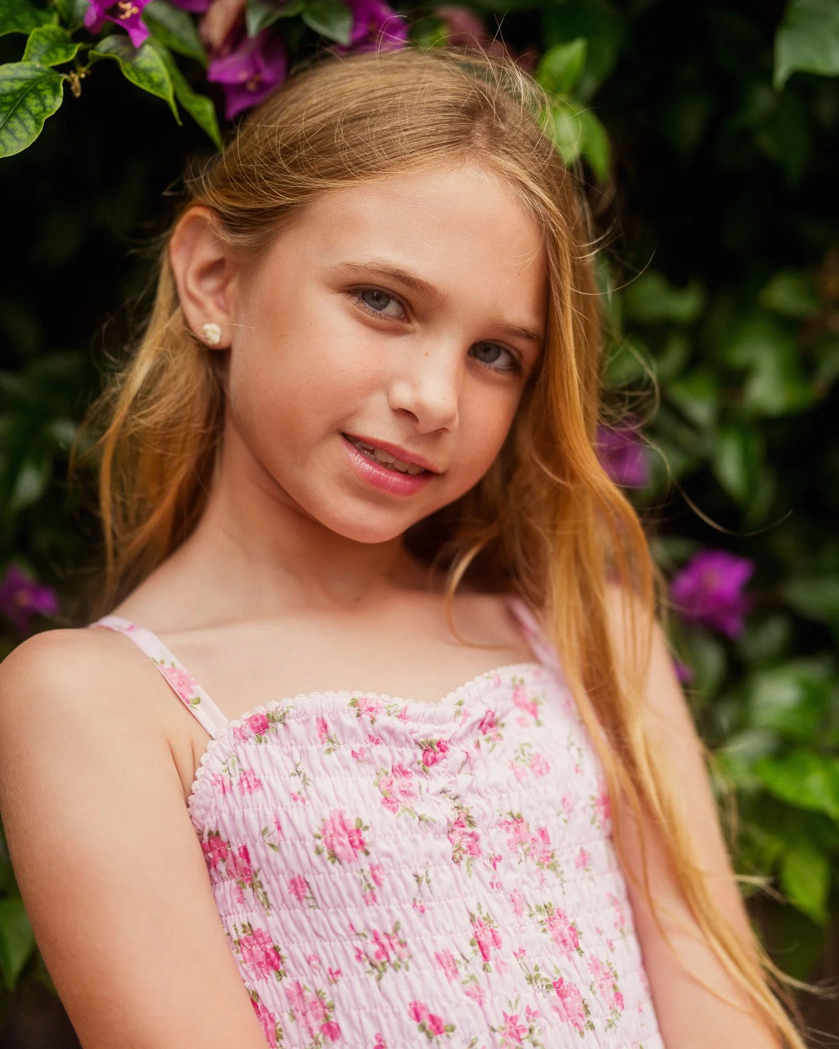 Close-up of a young girl with long strawberry blonde hair, wearing a pink floral dress, standing in front of green foliage and purple flowers.