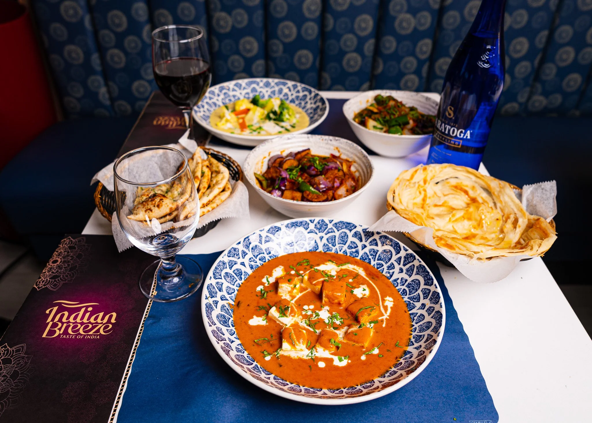 A table with Indian food including naan bread, curry, vegetable dishes, a bowl of gravy, and glasses of water and wine.