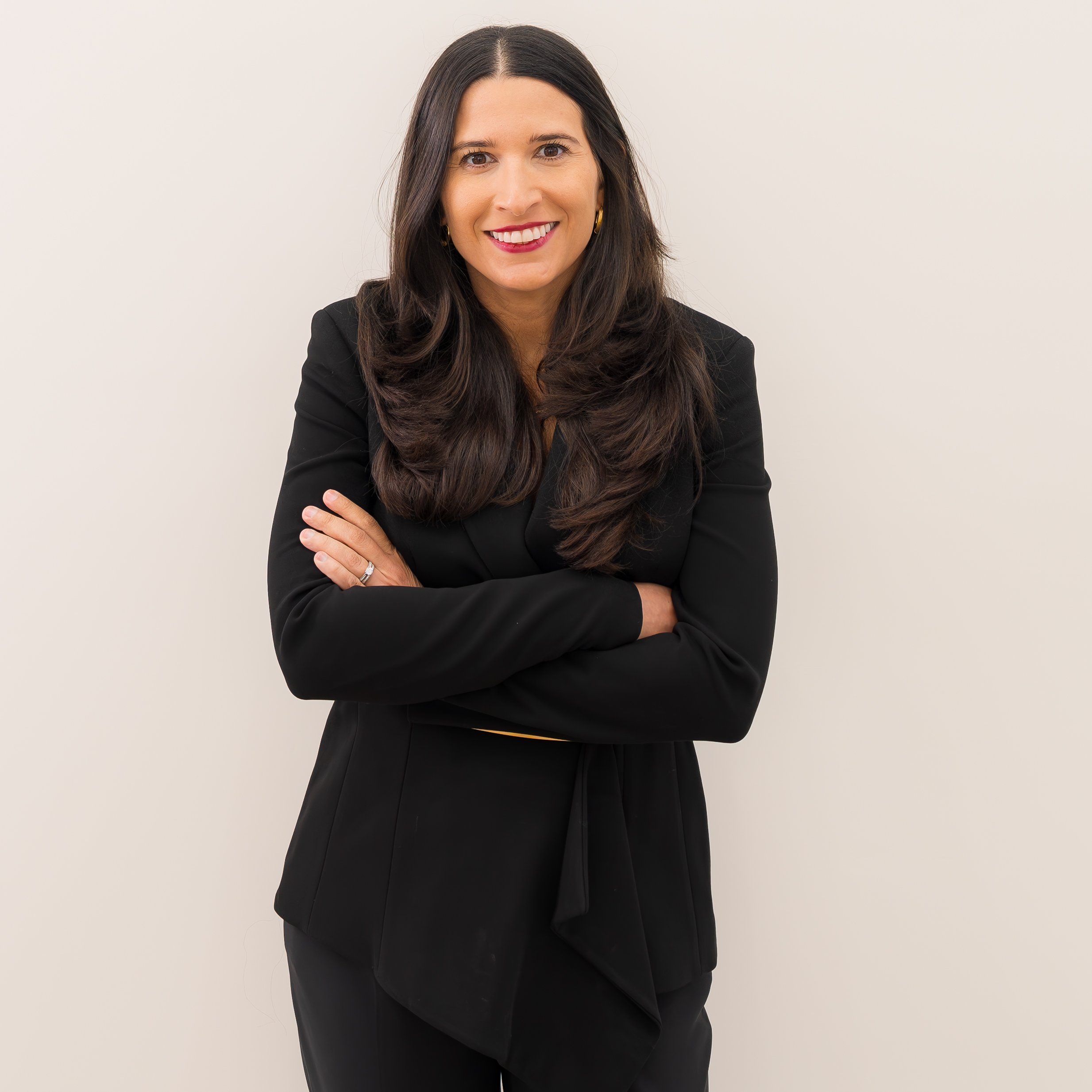 A woman with long dark hair, wearing a black blazer and smiling with arms crossed, standing against a plain white background.