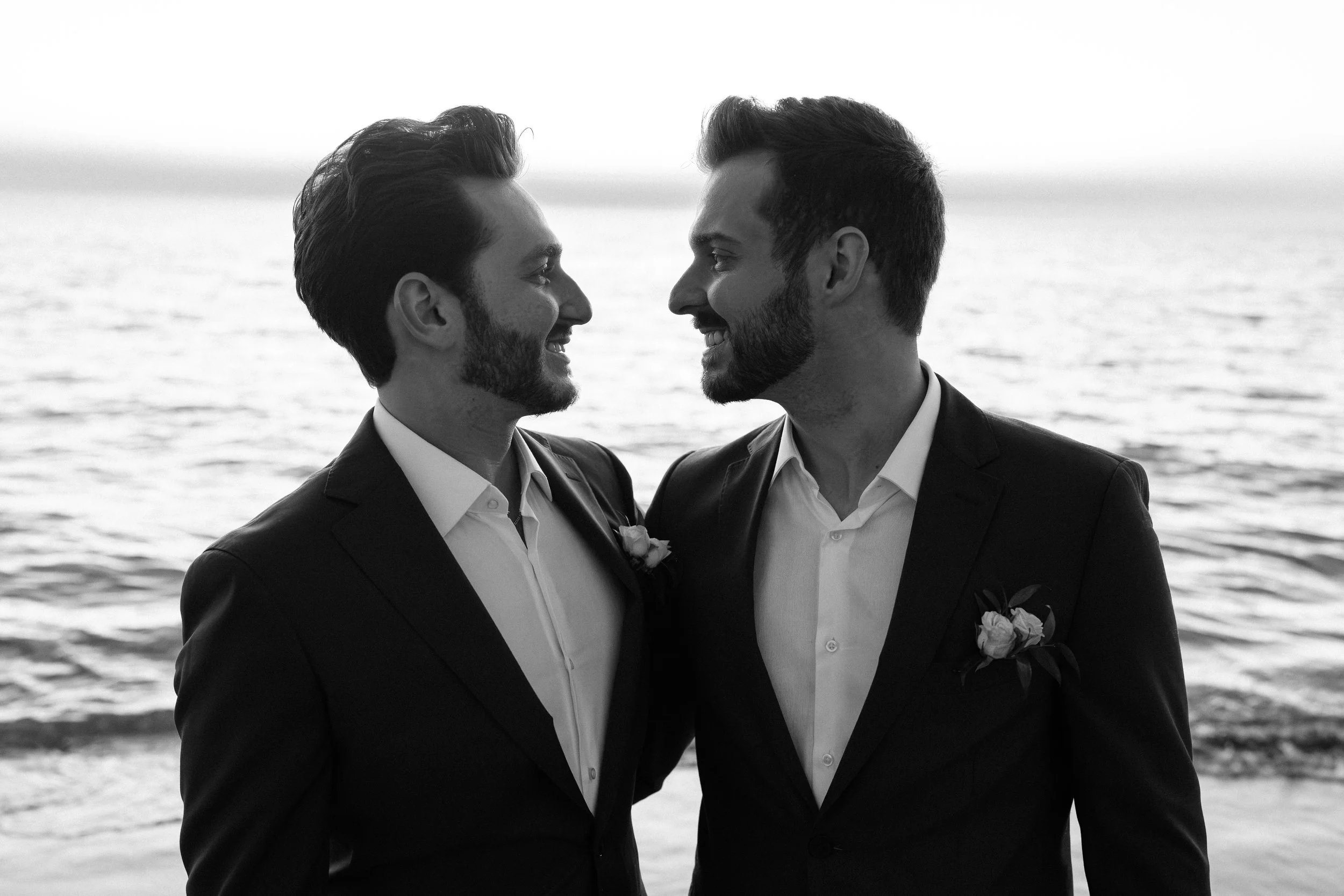 Two men in suits smiling at each other on the beach with ocean in the background, face to face, close to each other.