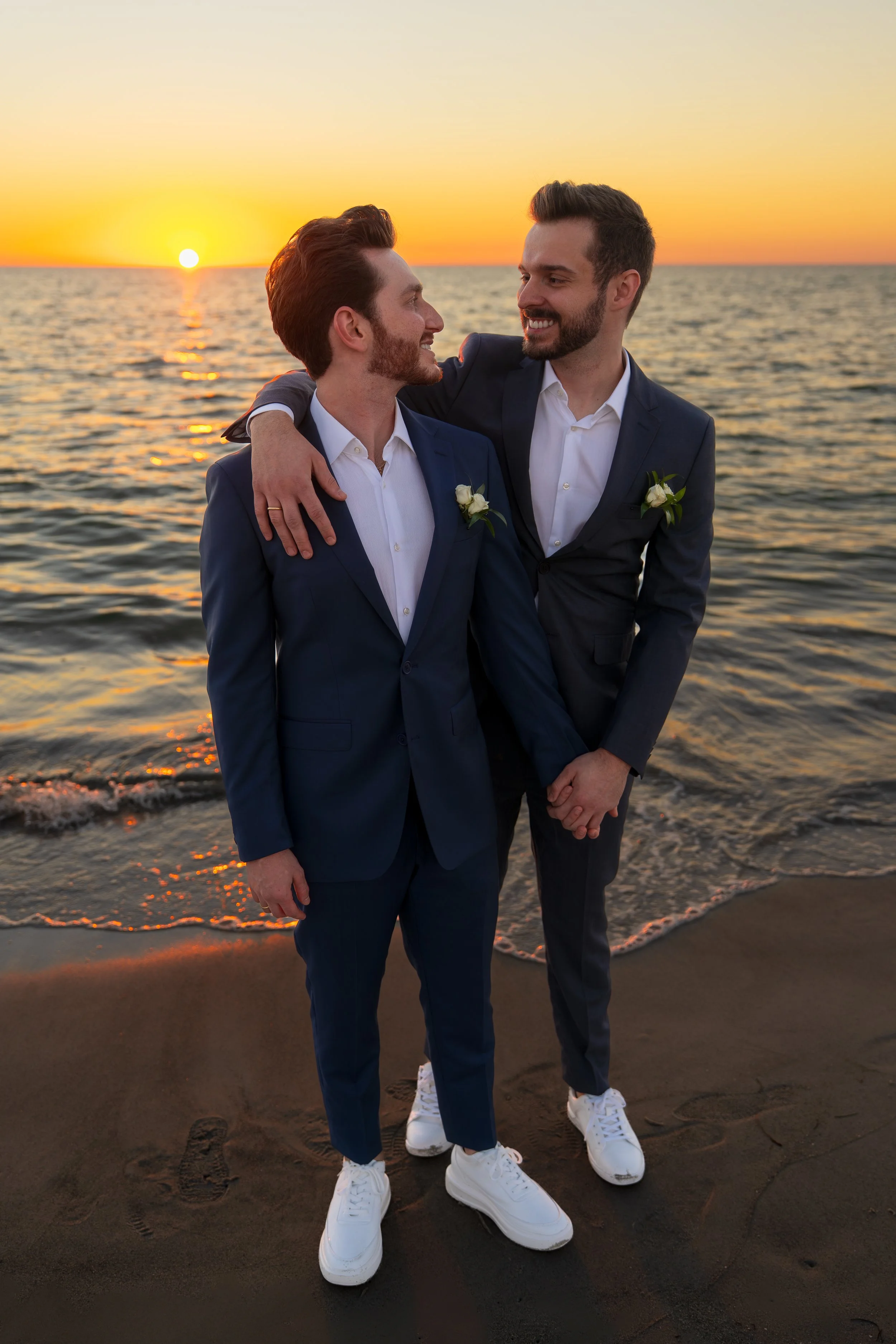 Two men in suits holding hands on the beach at sunset, with the ocean in the background.