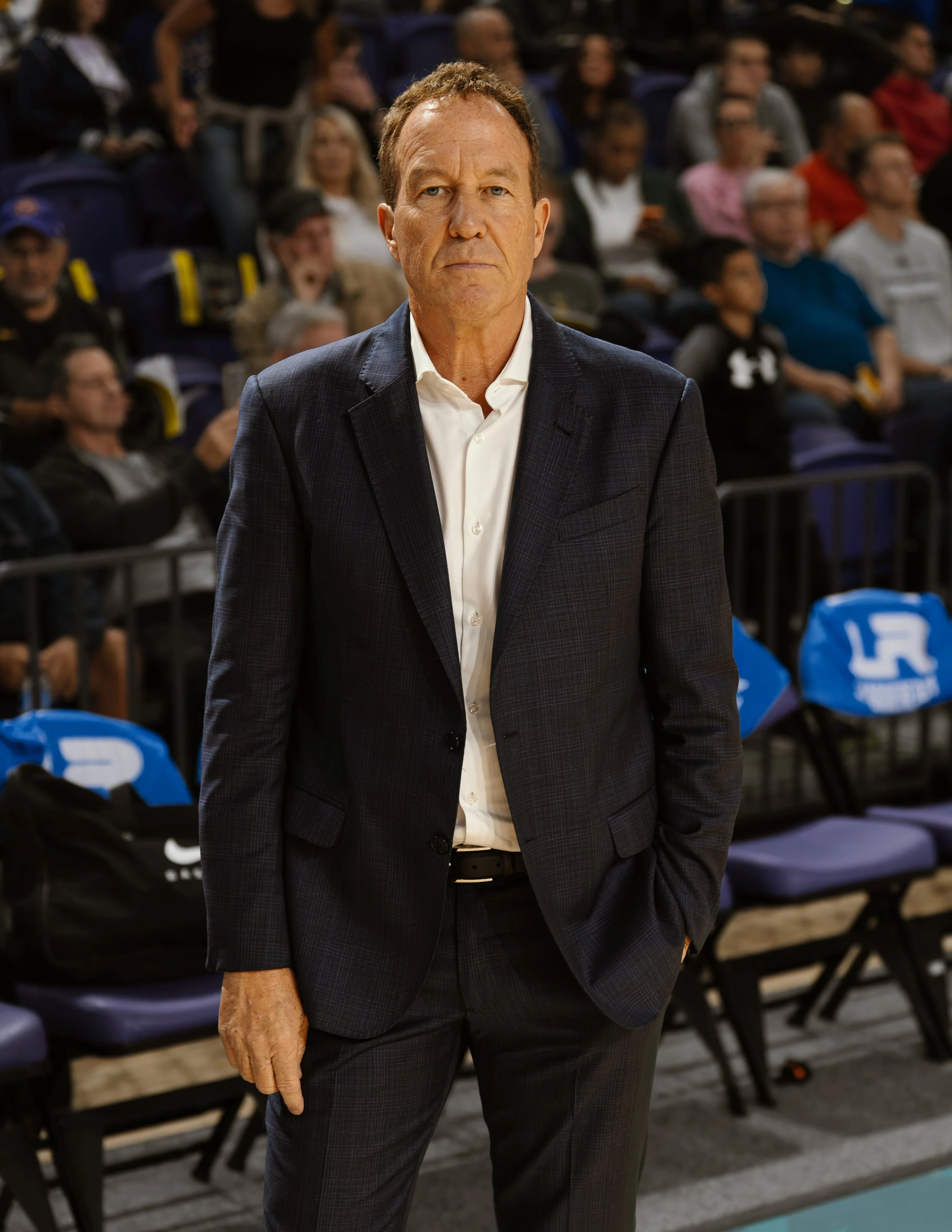 A man in a dark suit with a white shirt stands courtside at a sporting event, with spectators seated behind him.