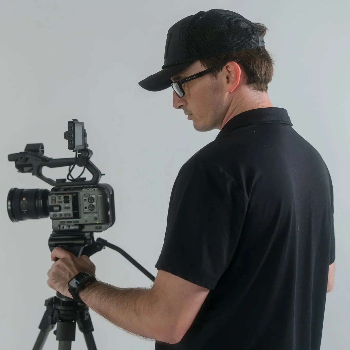 A man in a black cap and shirt operating a professional video camera mounted on a tripod.