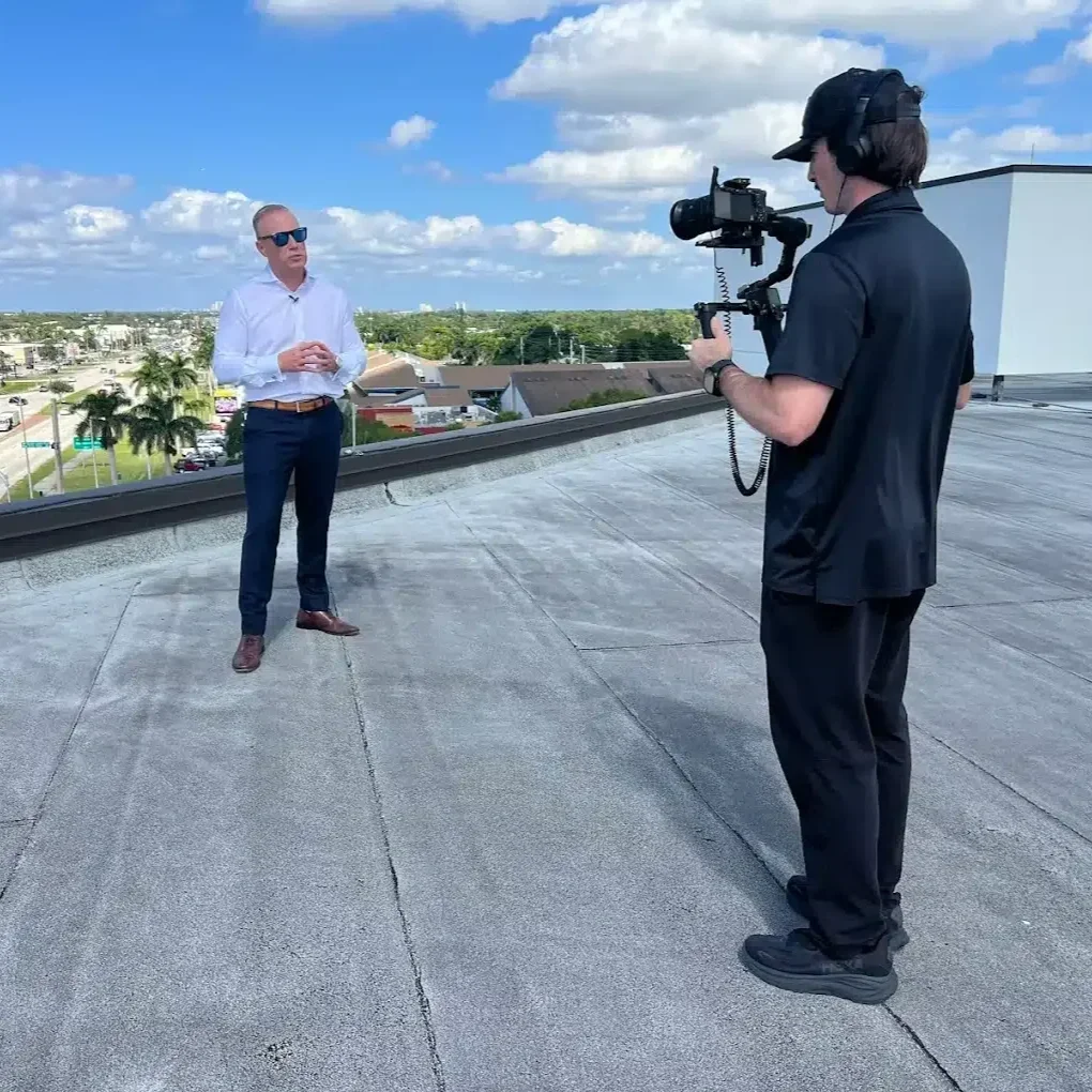 A man in sunglasses being interviewed on a rooftop by a videographer using a camera.