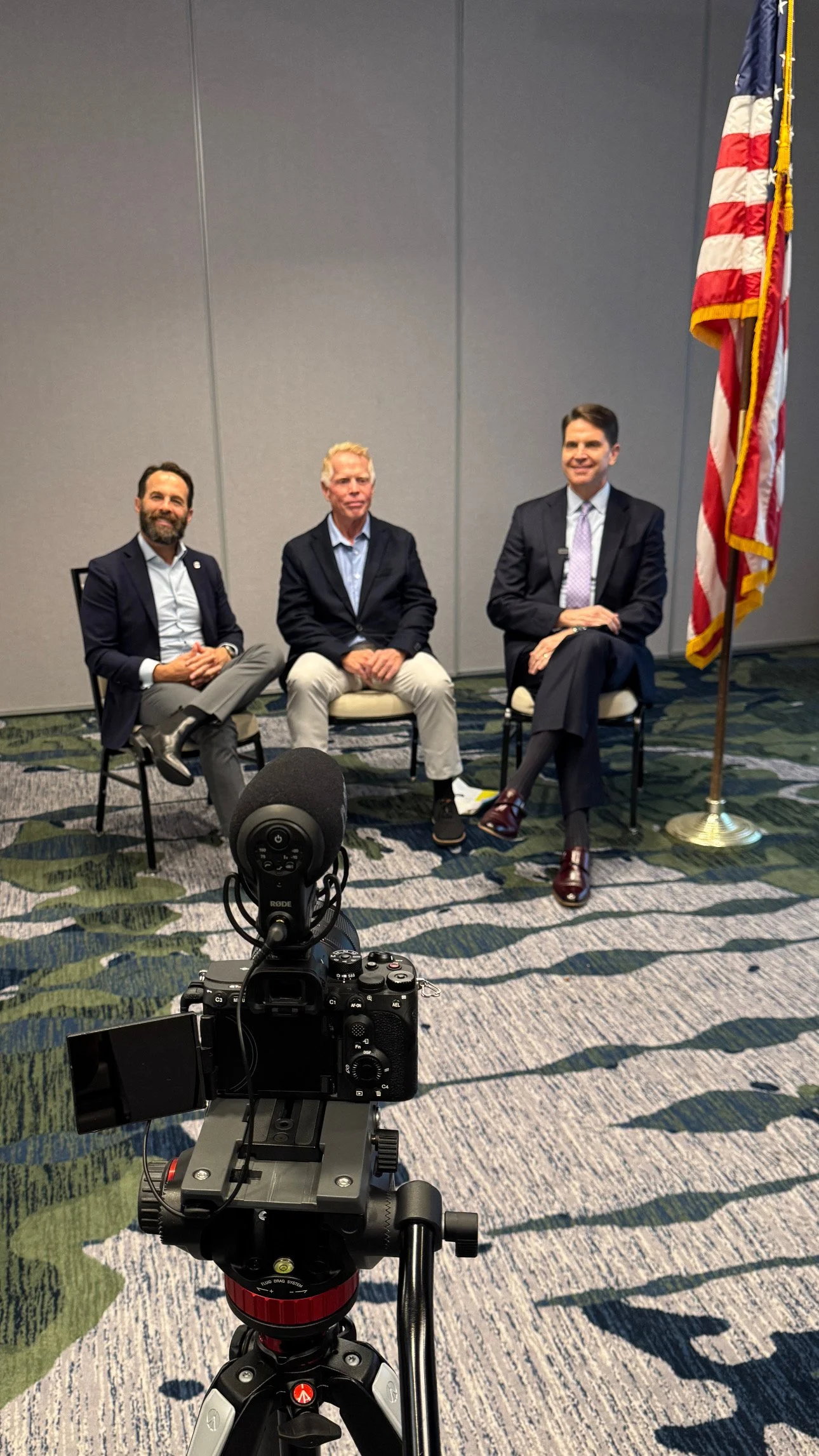 Three men sitting in chairs in front of a gray wall and an American flag, with a professional camera on a tripod in the foreground.