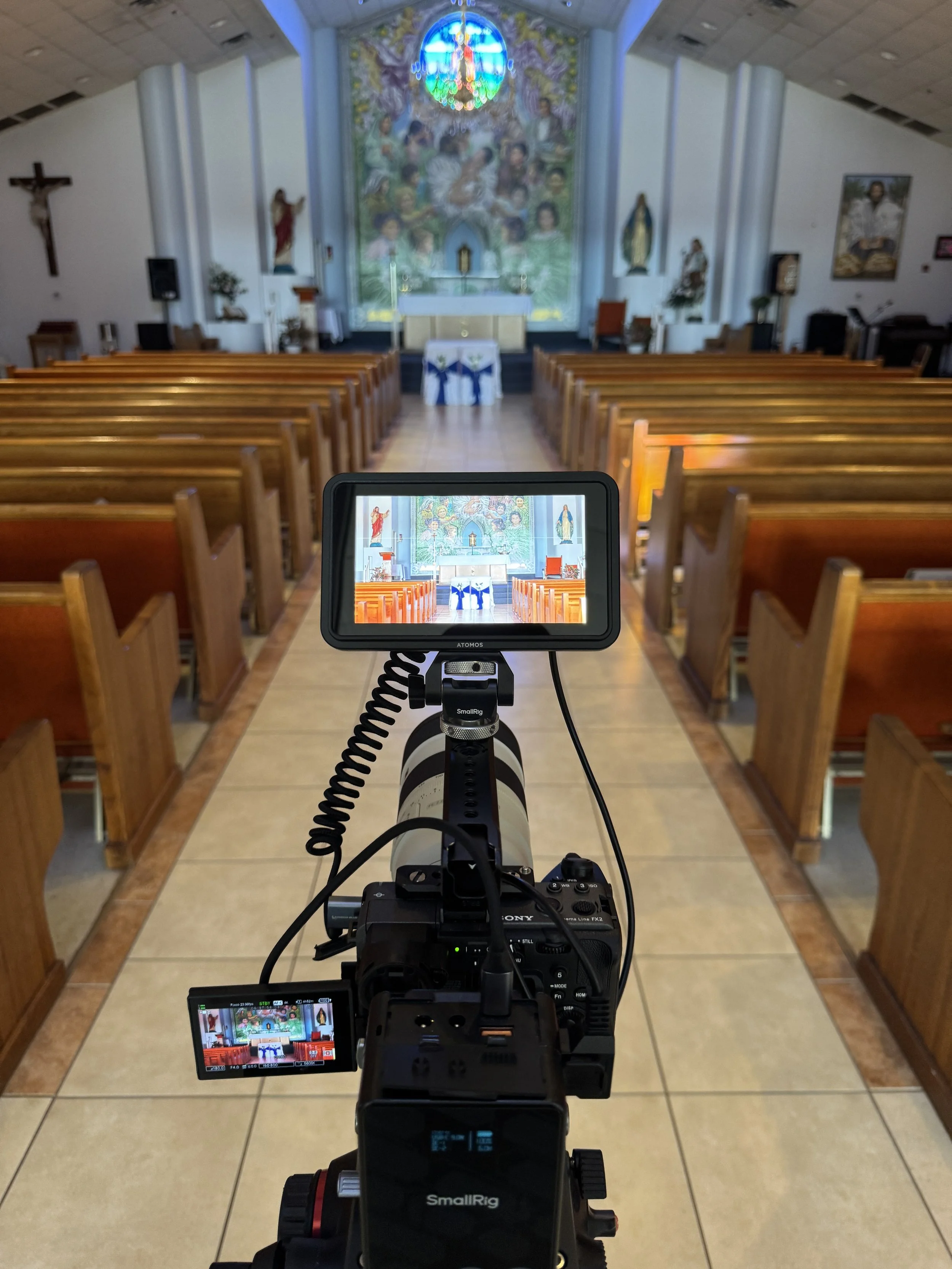 Interior of a church with wooden pews, altar, religious statues, crucifix, and stained glass window. The photo is taken through a camera viewfinder showing the church interior.