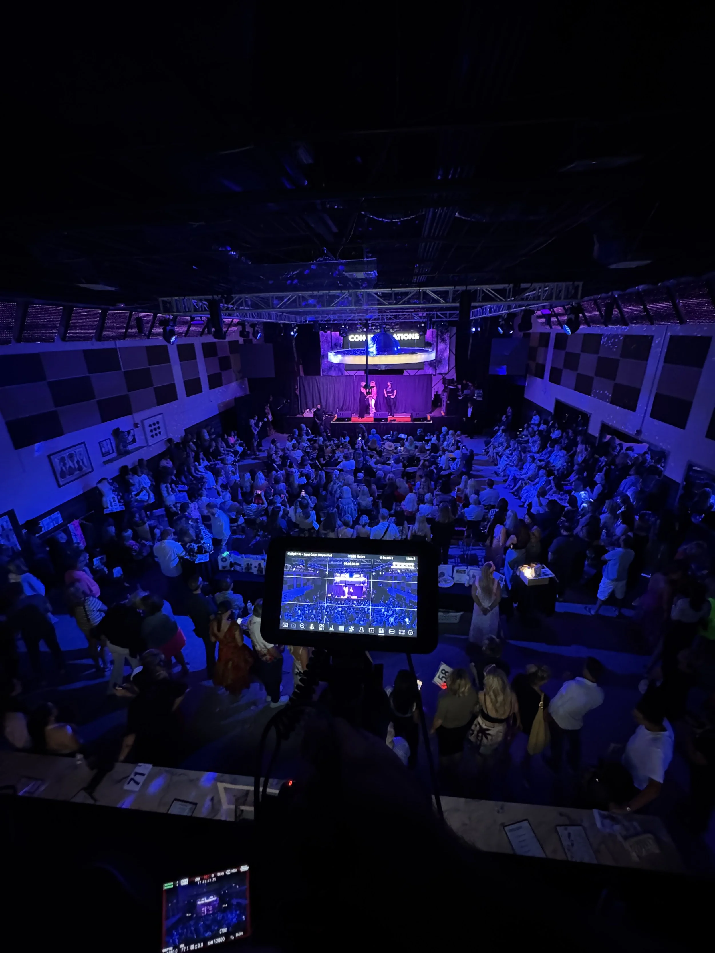 Concert stage with live performers under purple and blue lighting, audience seated and standing, event signage overhead, and camera screens in foreground capturing the scene.