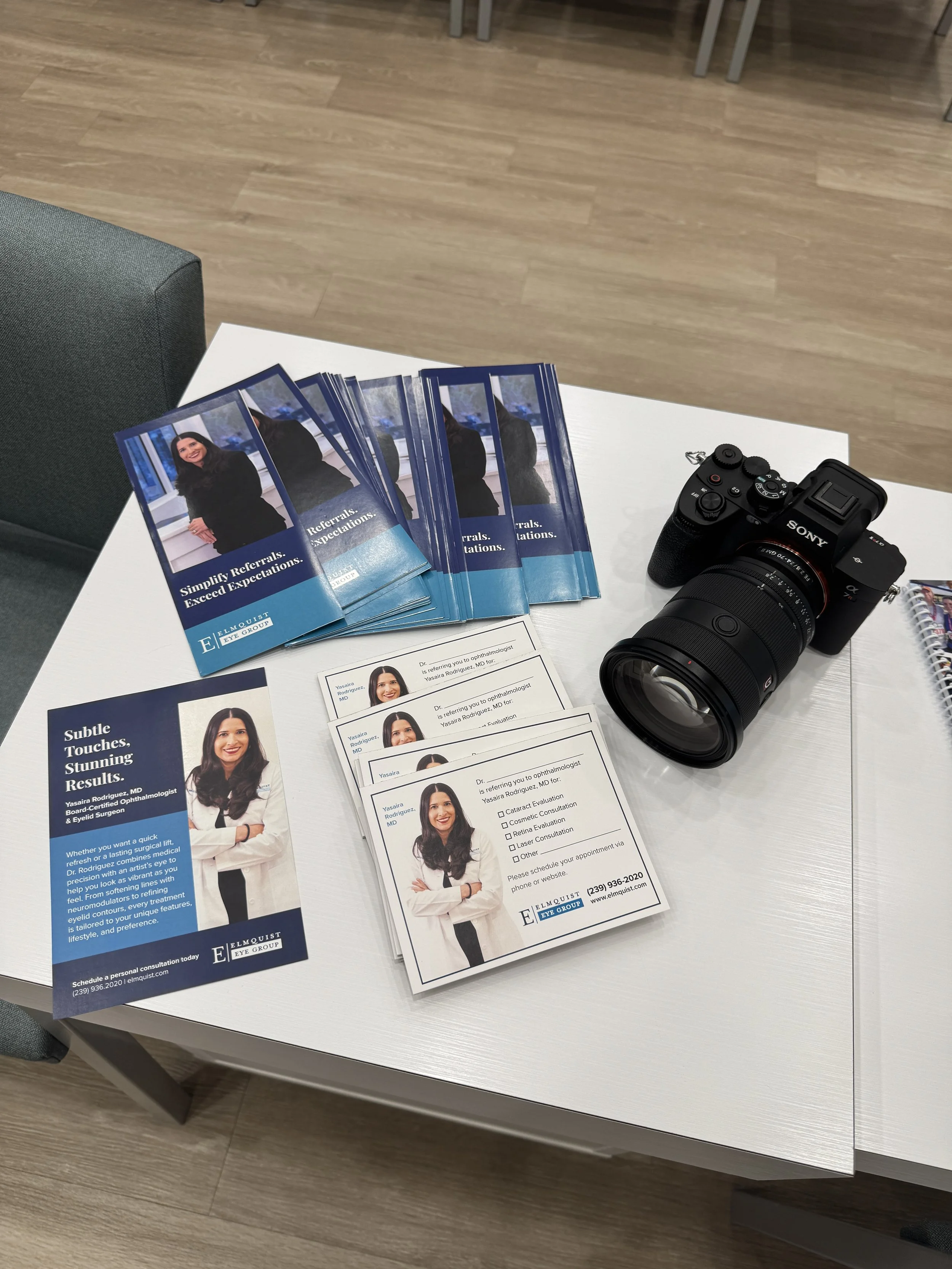 A white table with various brochures and pamphlets for an eye care provider, along with a professional camera, in a modern office setting with wood flooring.