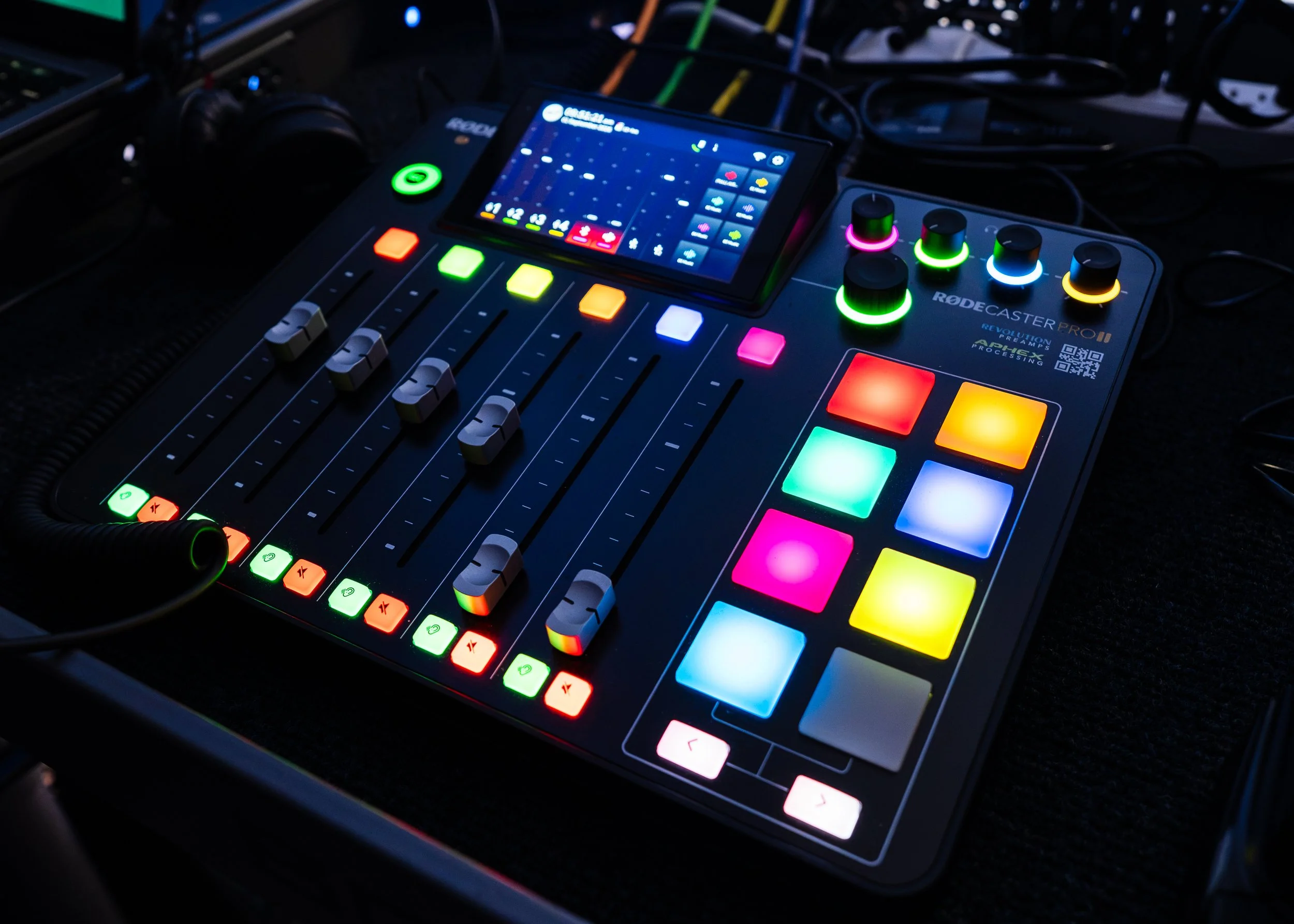 A professional DJ mixing console with colorful illuminated buttons and sliders, a small touchscreen display showing audio levels and controls, and headphones resting on the device, set in a dark environment.