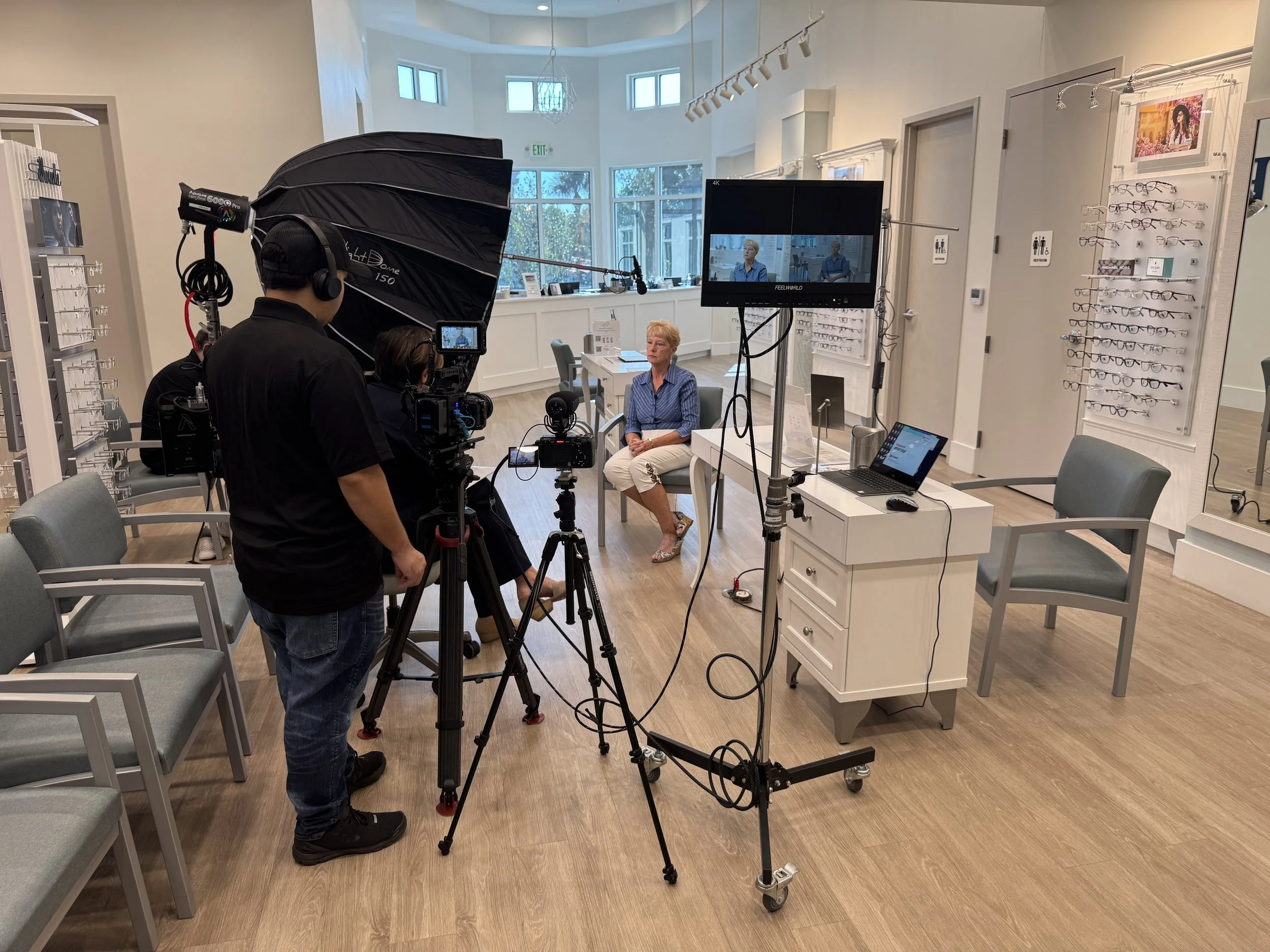 A film or television interview taking place in a well-lit room with large windows. An older woman with short blonde hair is seated on a chair, facing the camera, wearing a blue striped shirt and white pants. Two camera operators are filming her with 